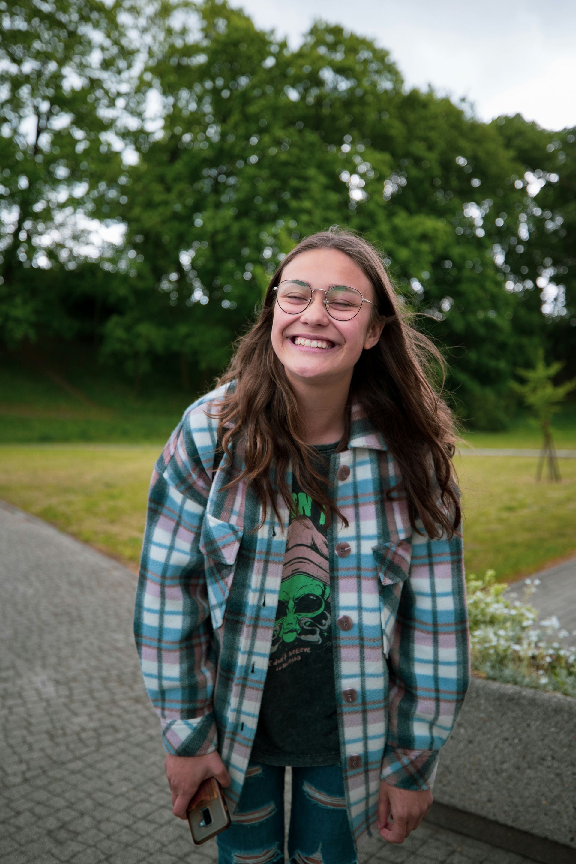 Person smiles broadly in a plaid shirt, holding phone, in a park setting.