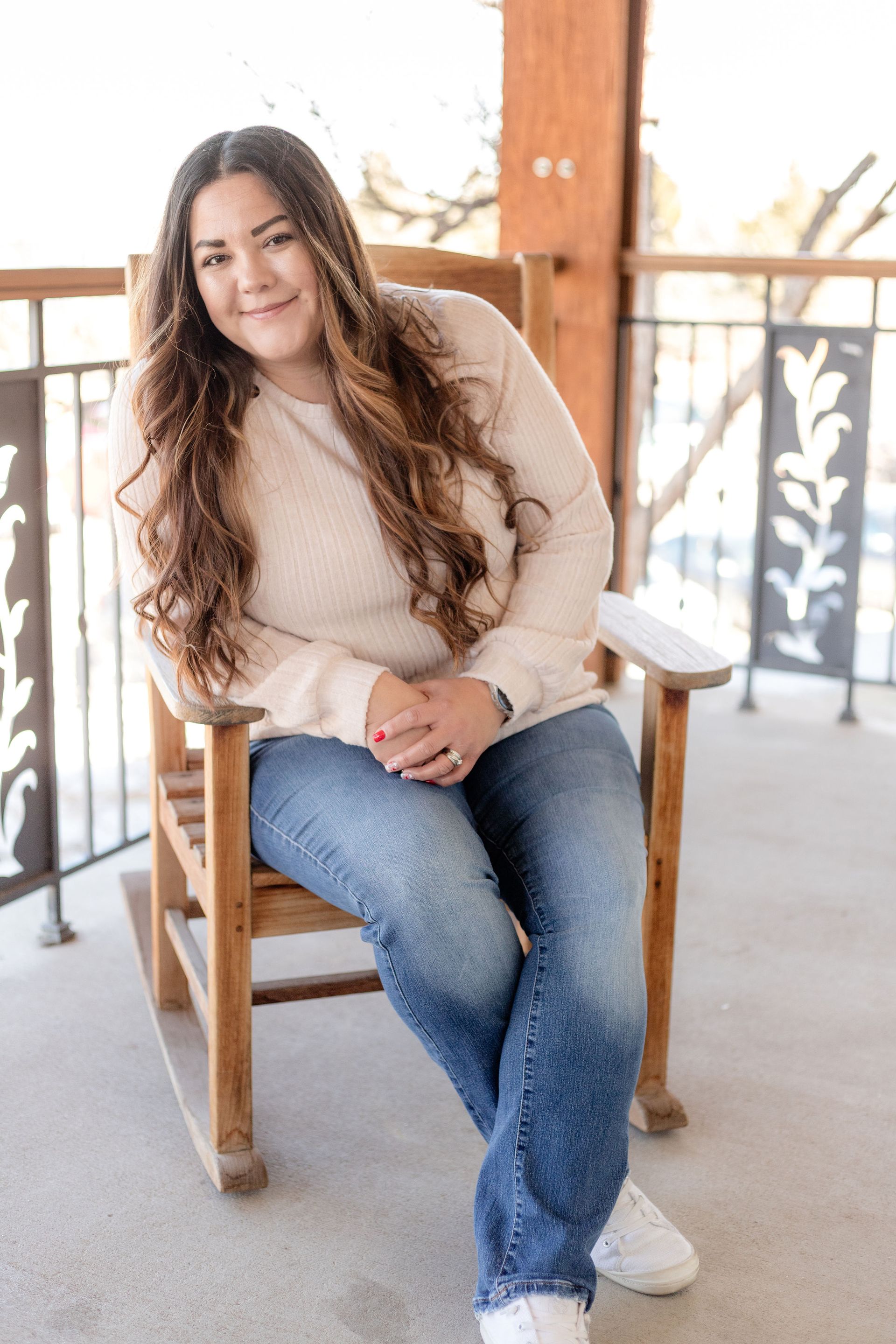 Woman sitting in a wooden chair on a porch, wearing a beige sweater and jeans, smiling at the camera.