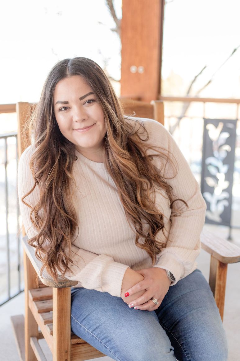 Woman sitting on a wooden chair, smiling in a beige sweater and jeans on a porch.
