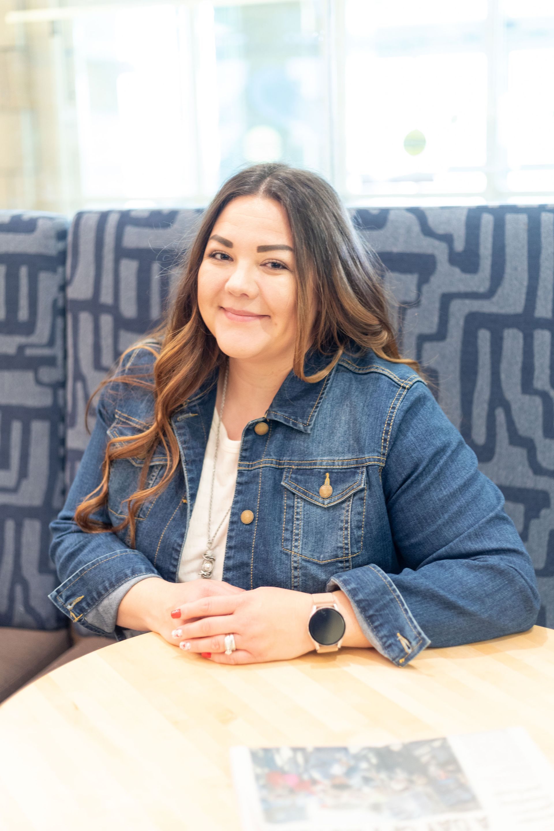 Woman sitting at a table in a denim jacket, smiling in a bright indoor lounge.