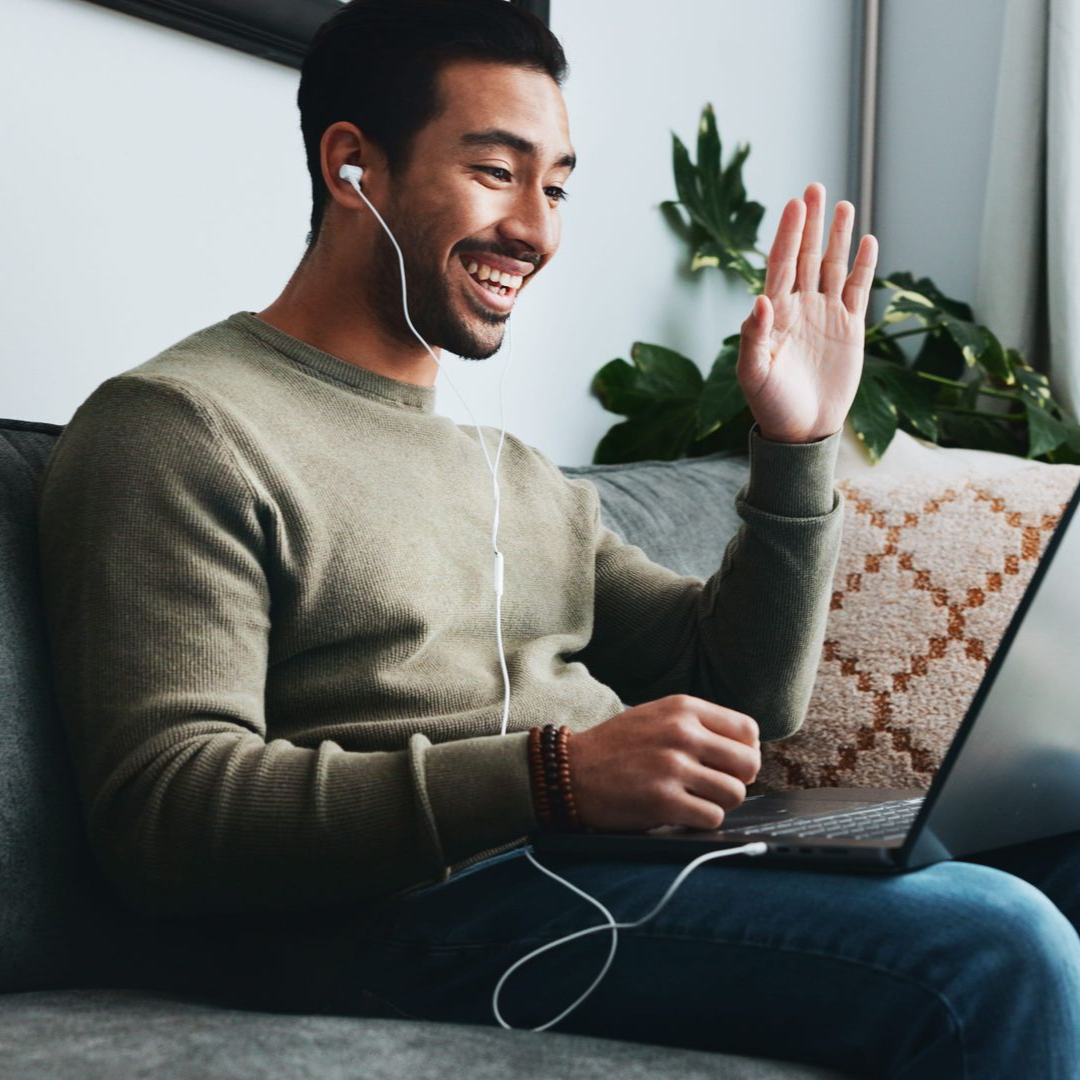Smiling man on a couch waving during a video call on a laptop, wearing earbuds.