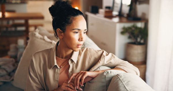 Woman with curly brown hair, wearing a brown sweater, gazes thoughtfully to the side. Sitting in a chair near a window.