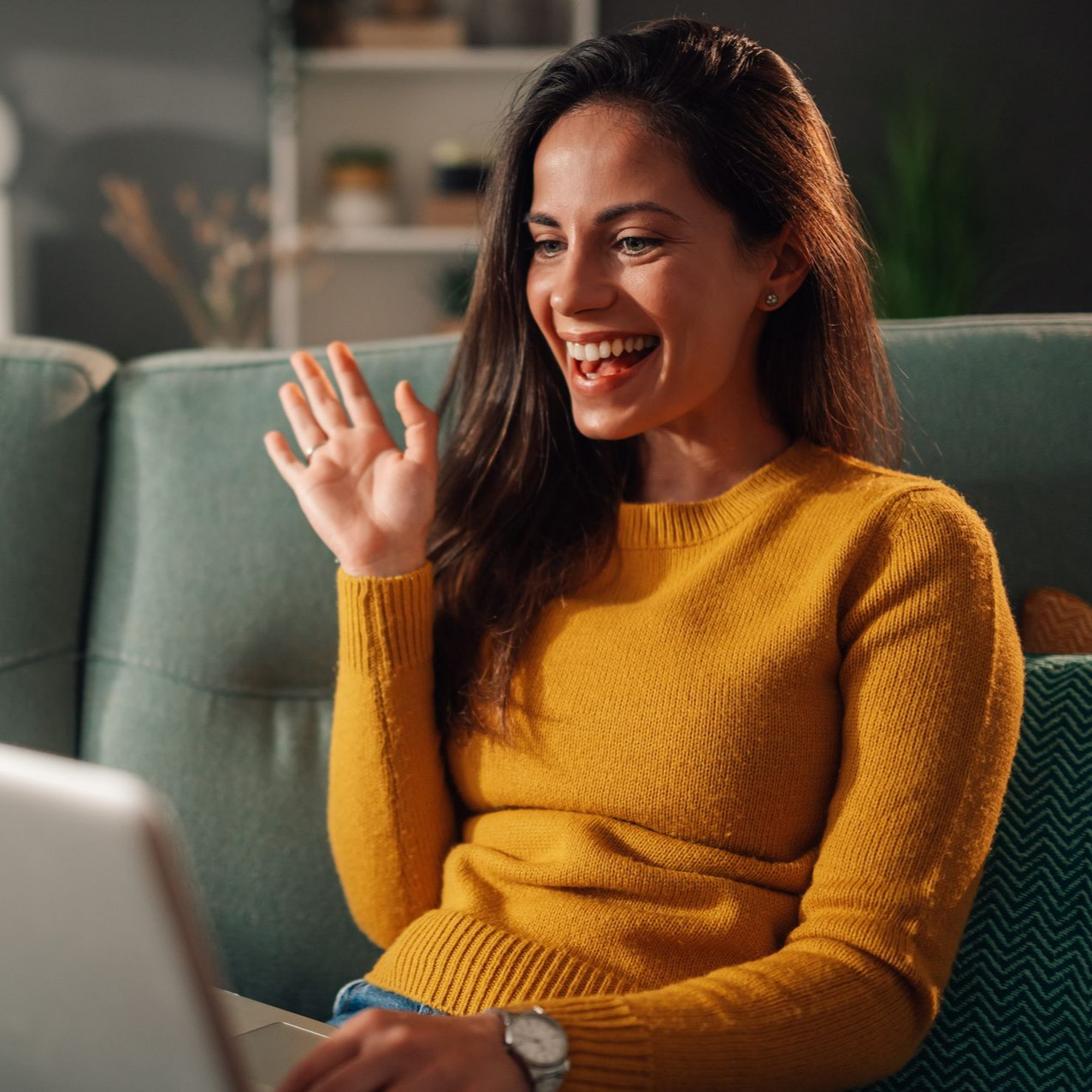 Smiling woman waving while sitting on a couch with a laptop in a cozy living room