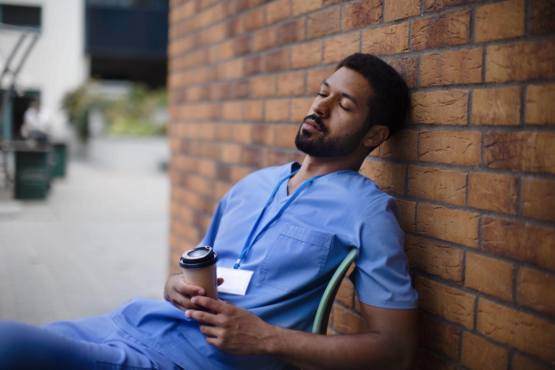 Man in blue scrubs leaning against a brick wall, resting with a coffee cup in hand.