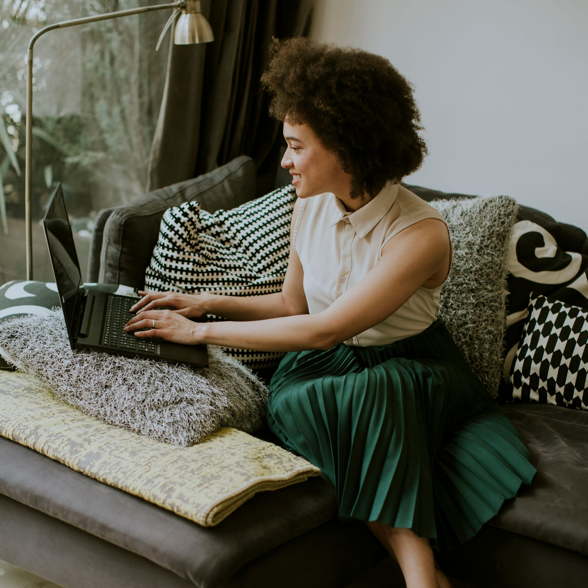 Person using a laptop on a couch with patterned pillows and a green skirt, in a cozy living room