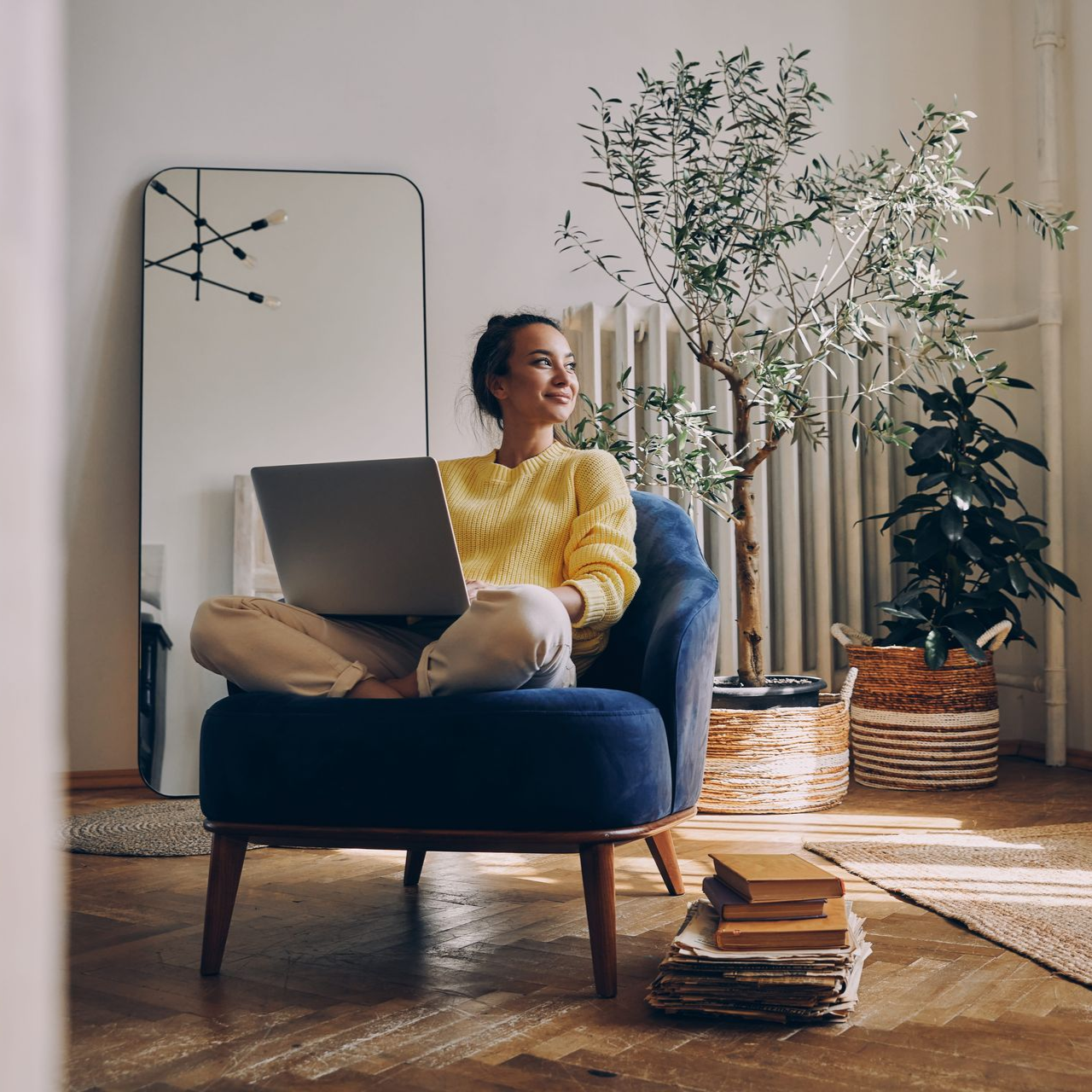 Person sitting on a blue chair with a laptop in a cozy living room beside plants and stacked books