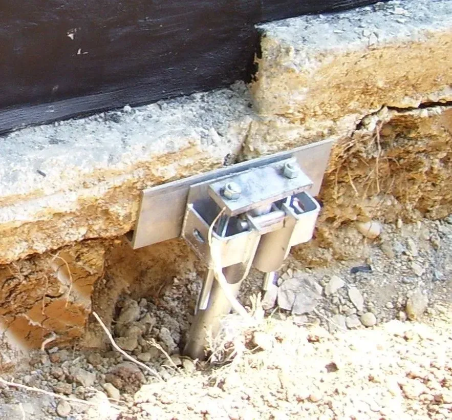 Metal bracket and ground rod anchor a structure's base, next to a weathered concrete foundation and exposed earth.