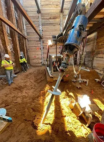 Construction workers operate machinery in a trench; a drilling machine is visible, along with brown soil.