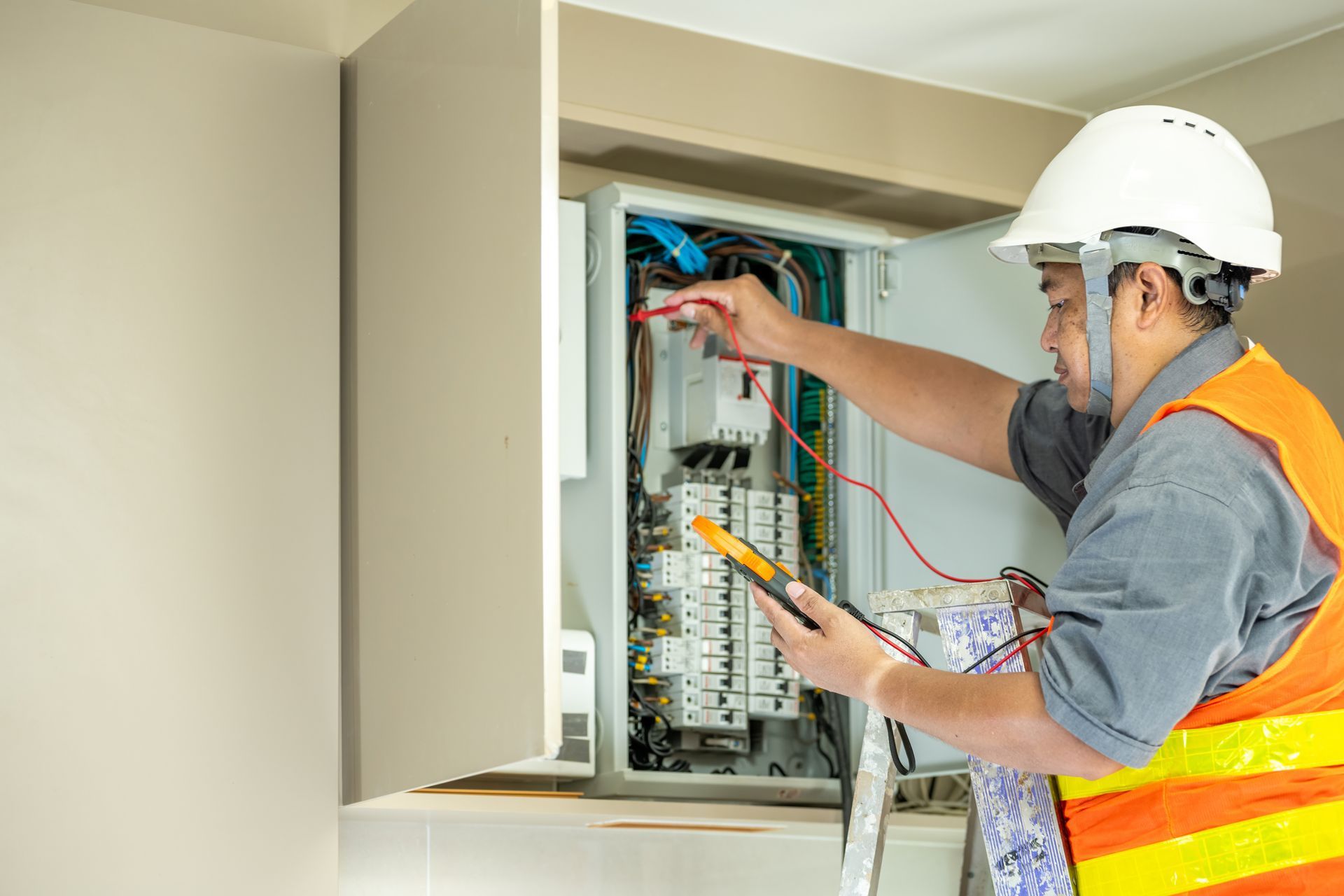 A commercial electrical contractor wears a helmet and a vest while working on an electrical panel.
