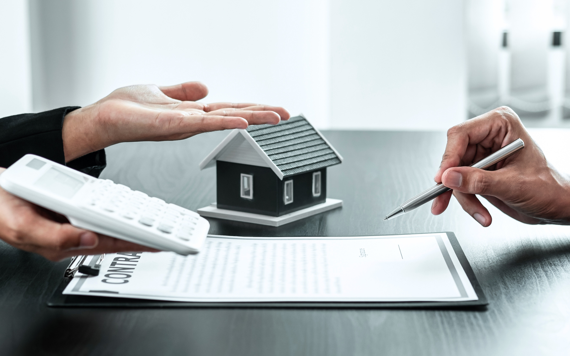 Hands and a pen review a house model and document on a desk during a property deal