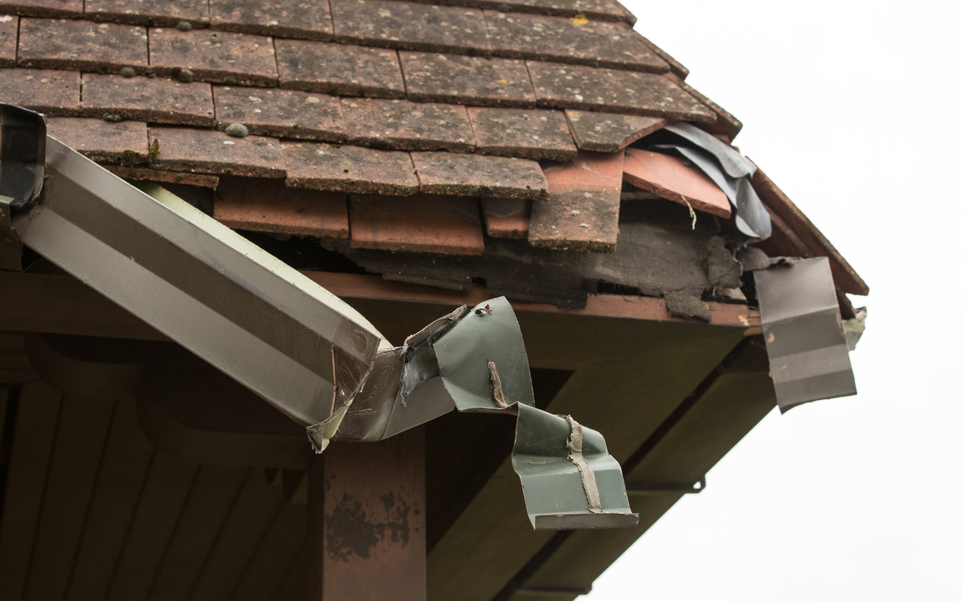 Damaged roof gutter hanging off brown shingled house roof with loose, bent metal pieces
