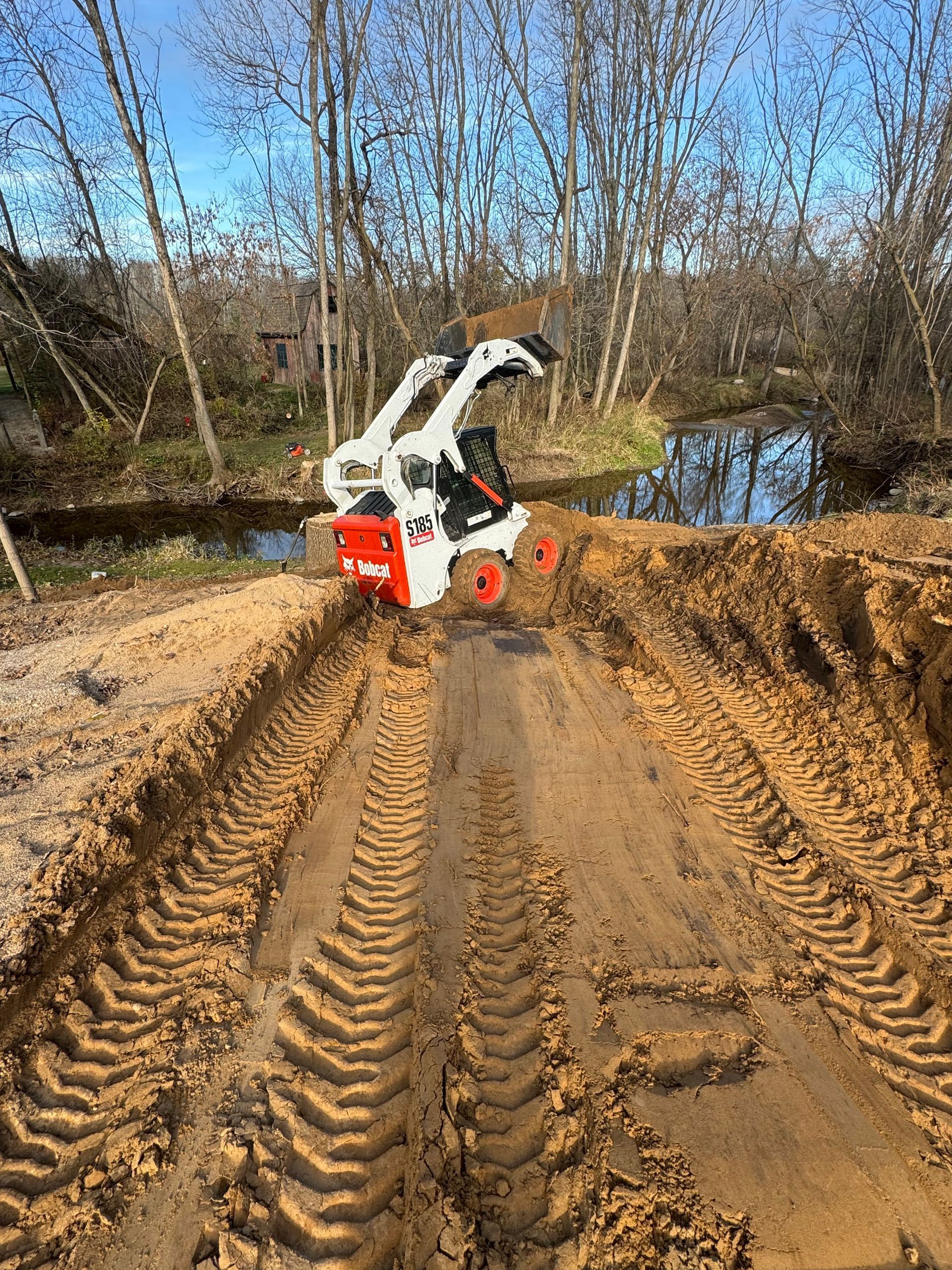 Small white-and-red excavator digging on a muddy trail beside a creek in a wooded area