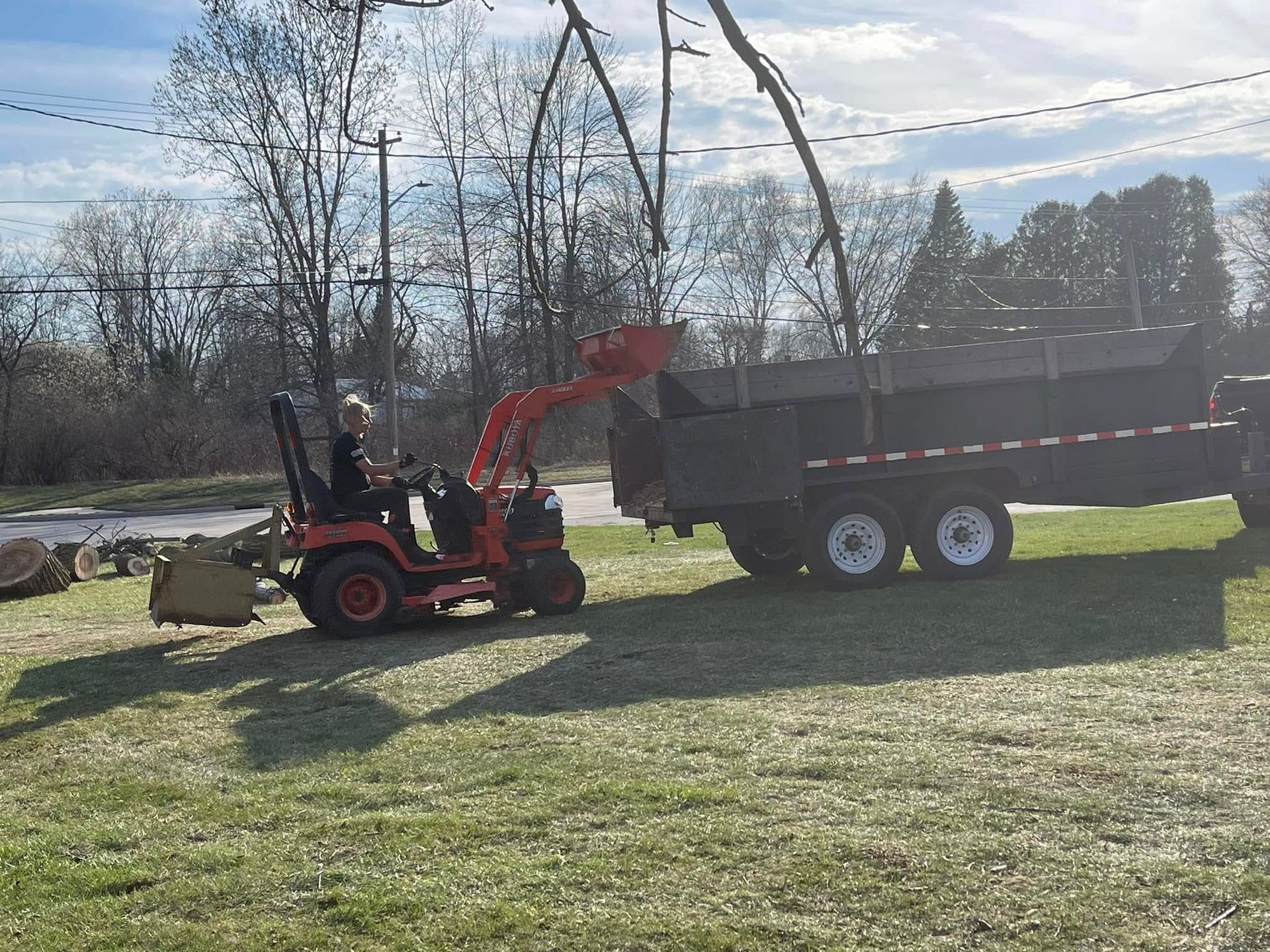 Small red loader lifting dirt into a dark dump trailer on a grassy field under a cloudy sky.