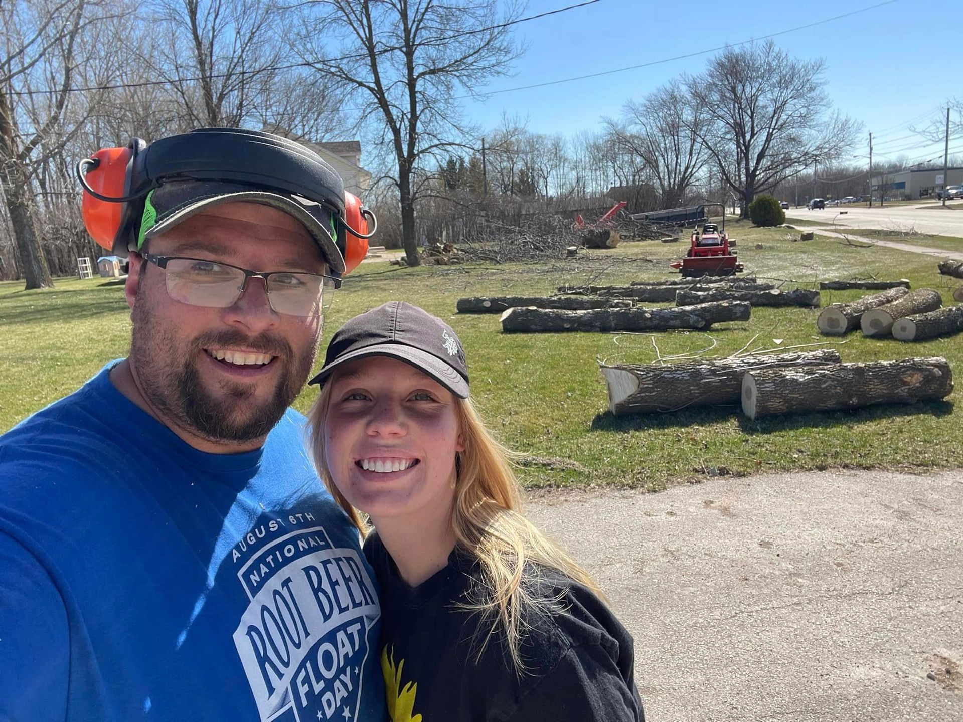 Two smiling people taking a selfie outdoors near stacked logs on a sunny day