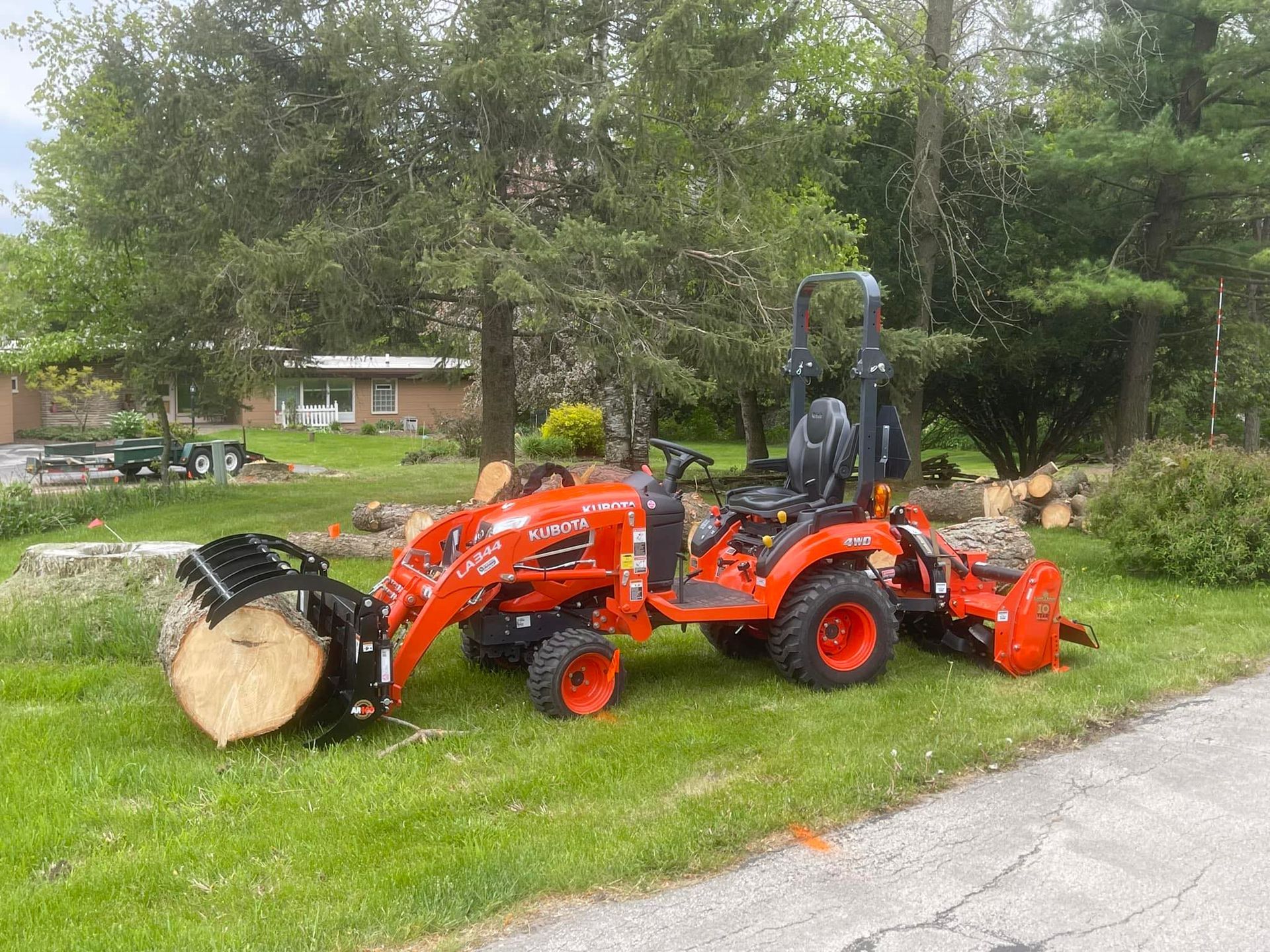 Orange tractor with front loader and mower attachment parked on a grassy lawn near a road