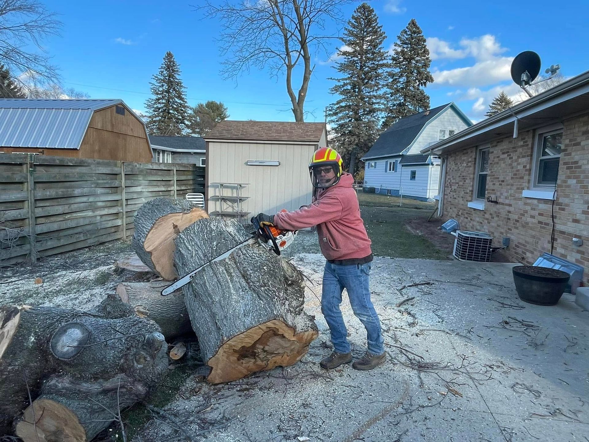 Person in red jacket and helmet pushing a large log in a snowy yard with piles of cut wood.