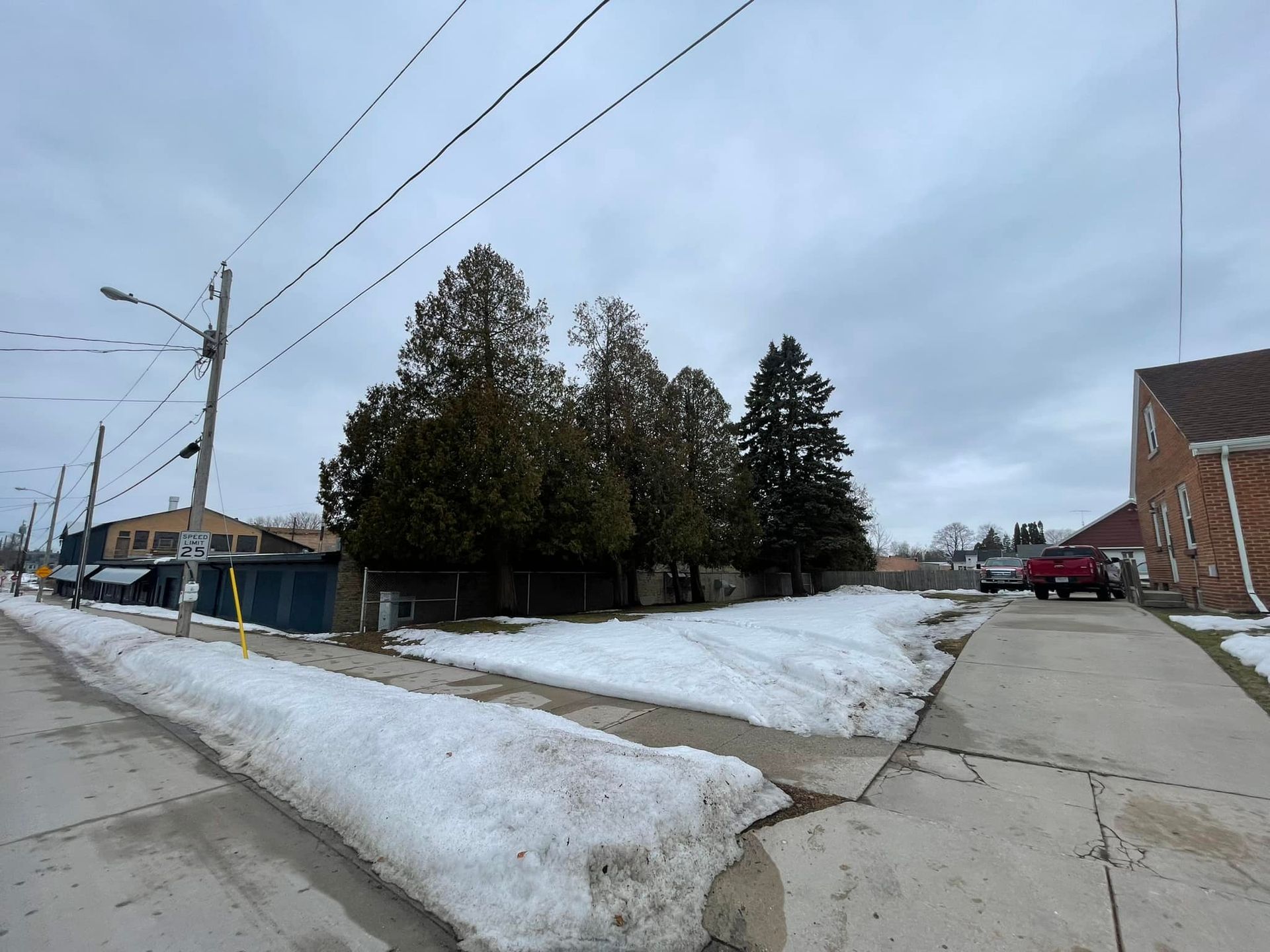 Snowy roadside with sidewalk, trees, utility poles, and a brick building under a cloudy sky.