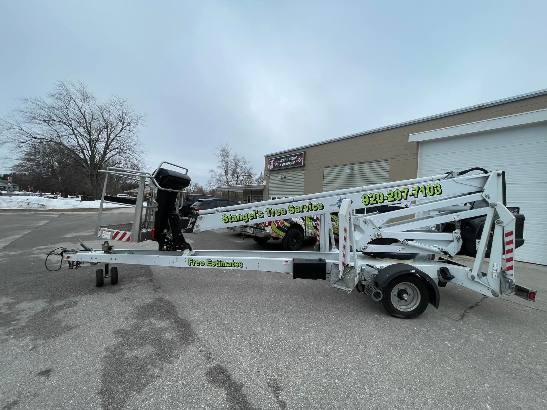 Small white utility trailer with folded boom lift parked outside a building in a snowy lot