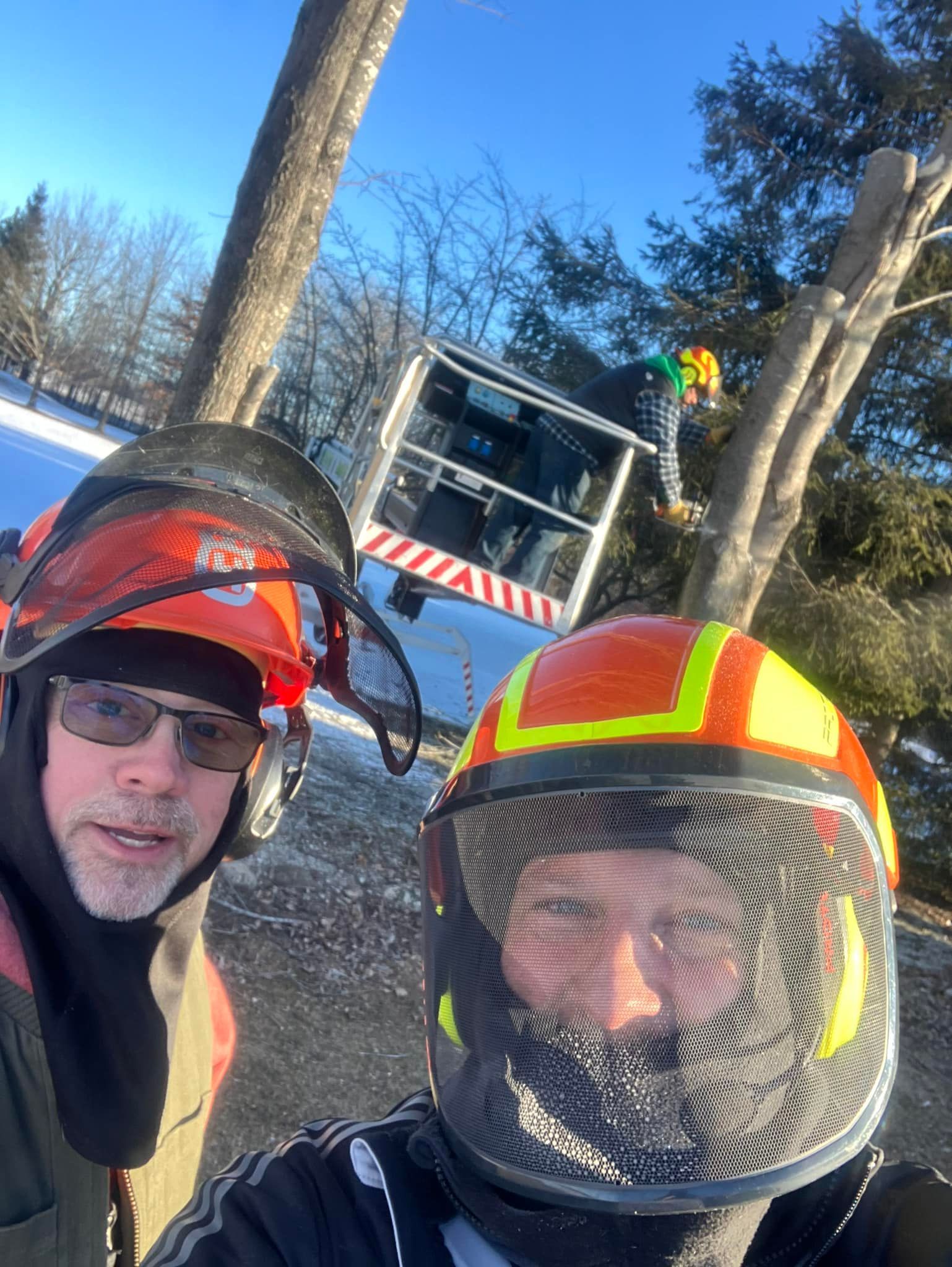 Two people in safety helmets posing outdoors near trees and a construction or work site