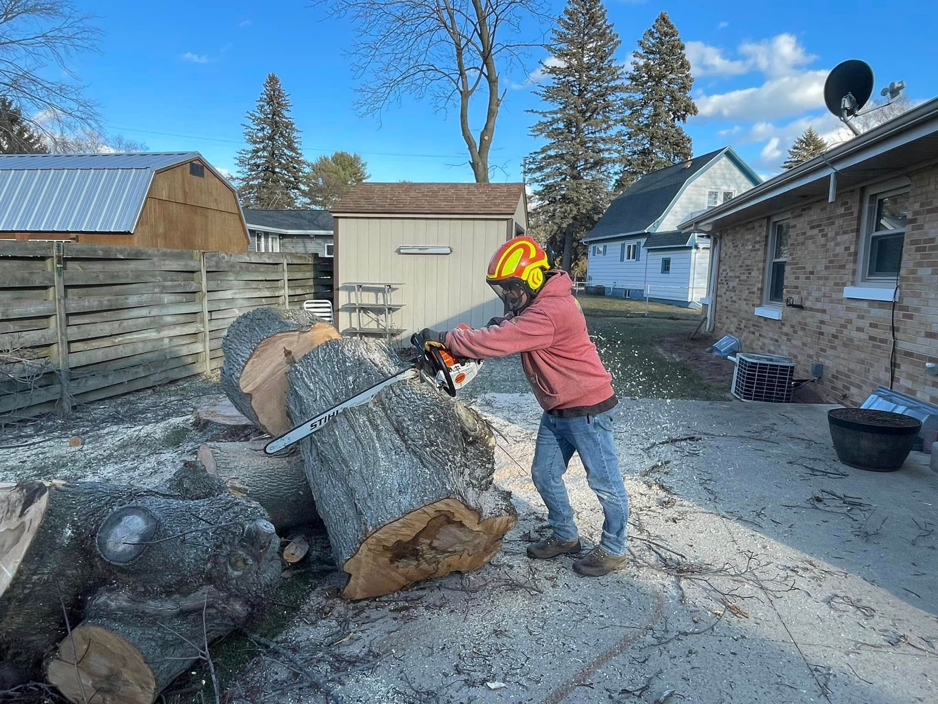 Person in red jacket cutting a large log outdoors with a chainsaw near a house on a sunny day