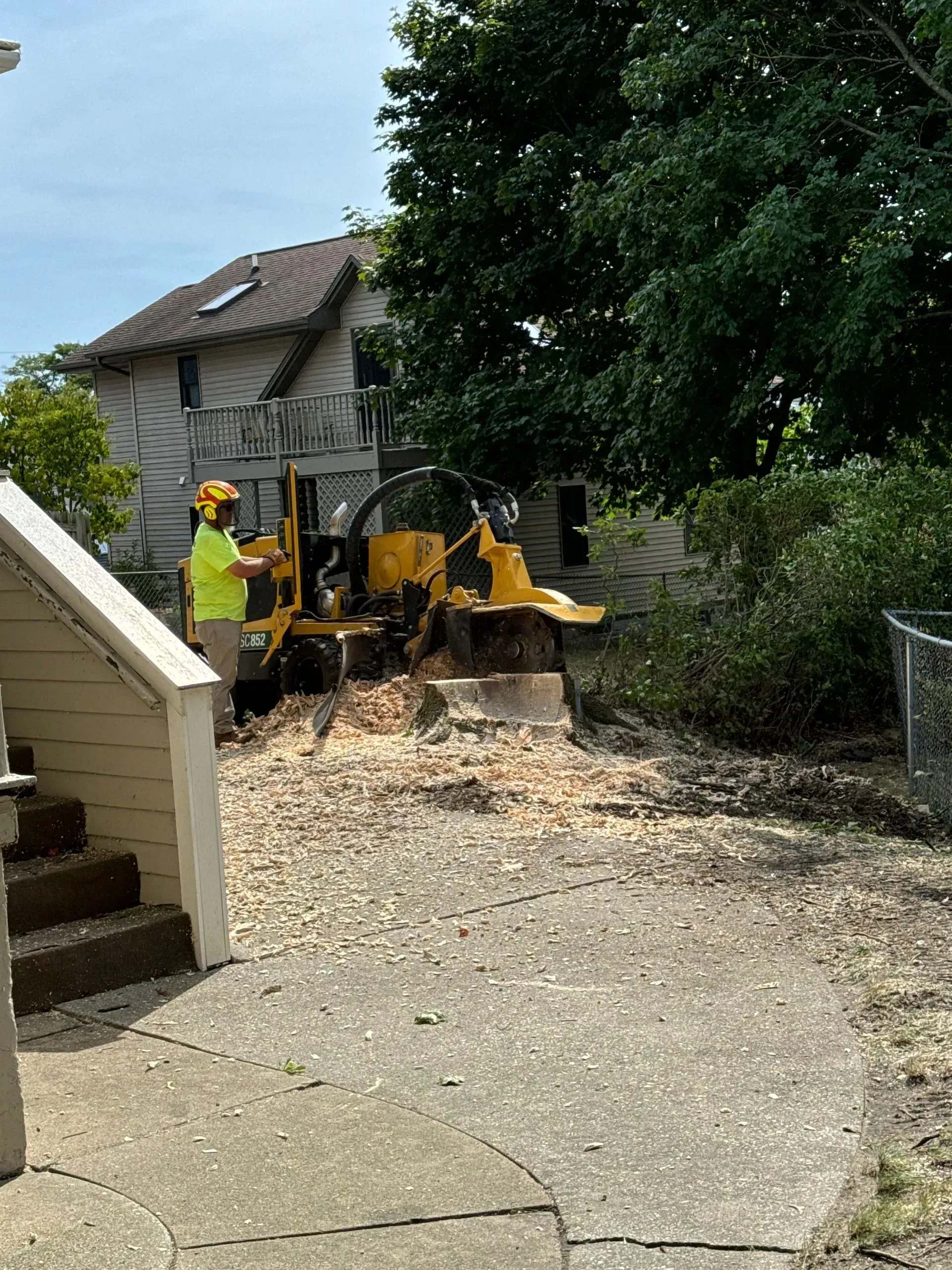 Roadwork crew with yellow excavator clearing debris in a driveway beside a house and tree
