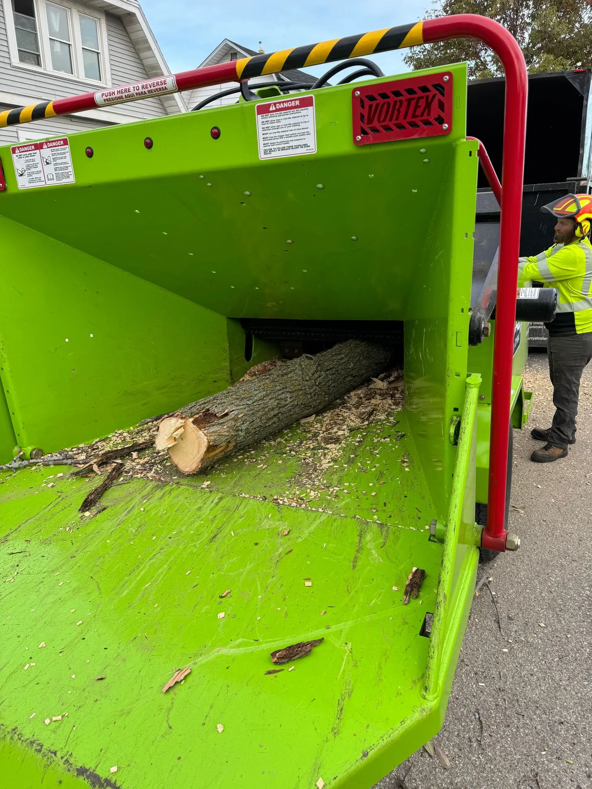 Green wood chipper with a log being fed in, worker standing beside it on a street.