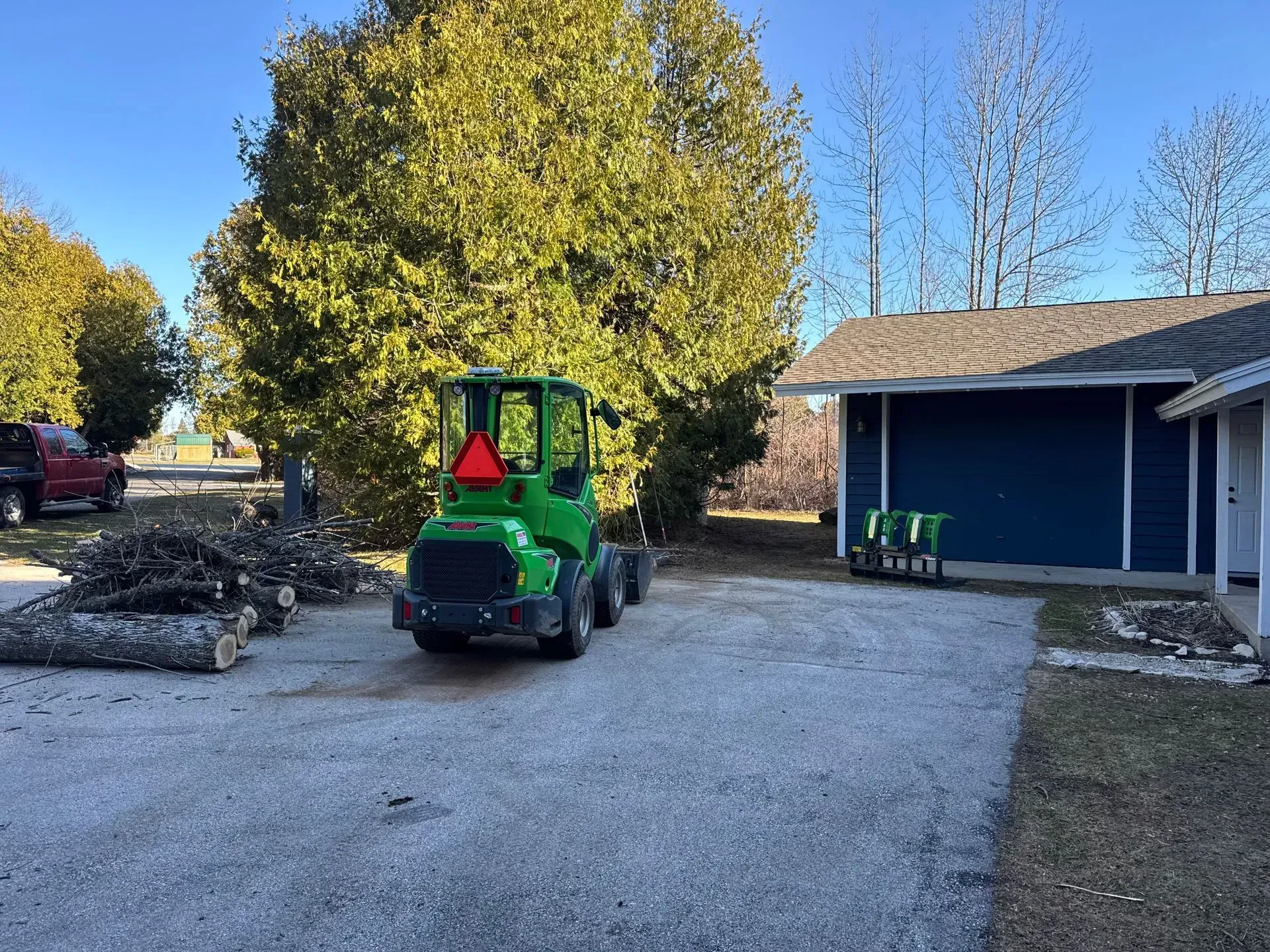 Green compact tractor in a gravel driveway beside a blue house, with a large tree and woodpile nearby.