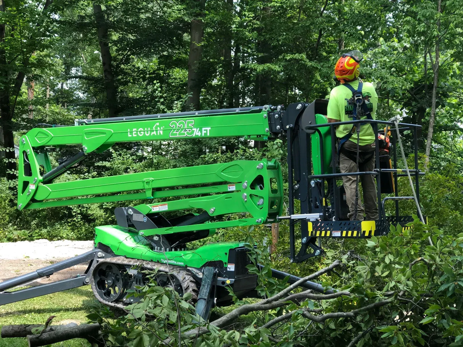 Green boom lift parked by roadside with worker in orange helmet beside trees