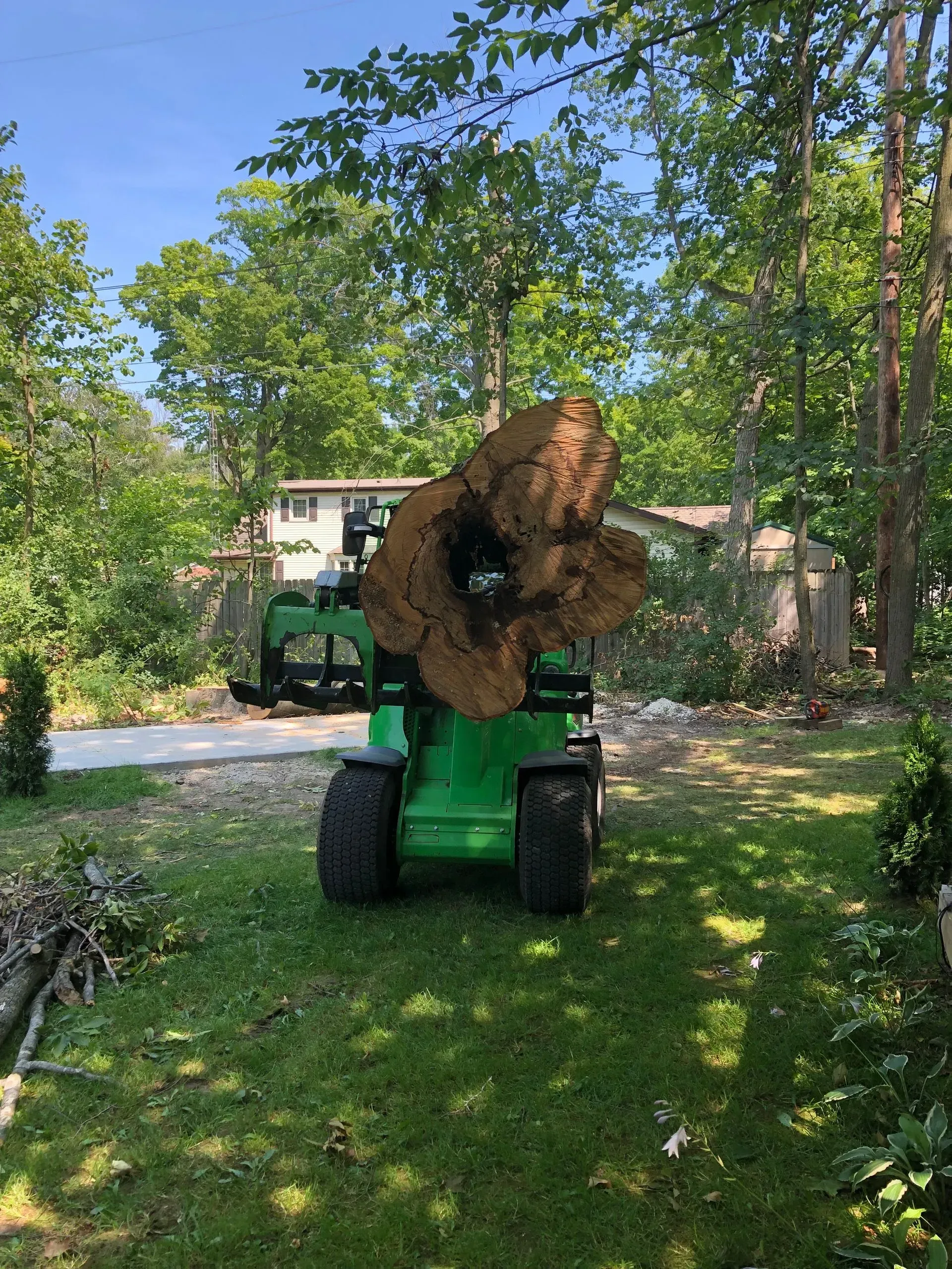 Person on a green tractor lifting a large brown bale in a sunny wooded yard