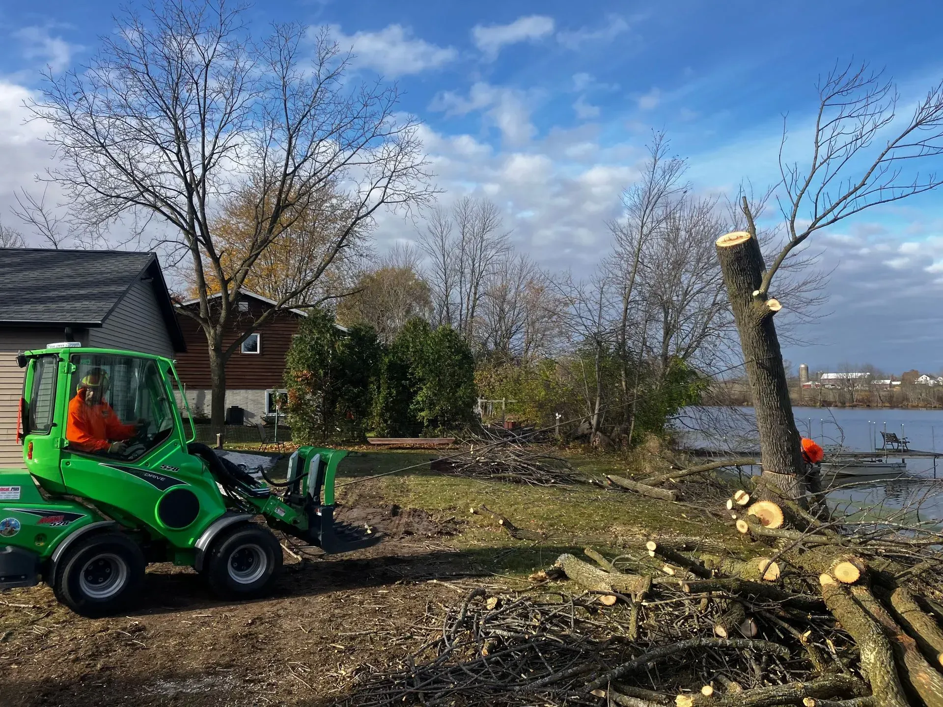 Green skid-steer clearing a yard with a cut tree trunk and branches near a lakeside house under a blue sky
