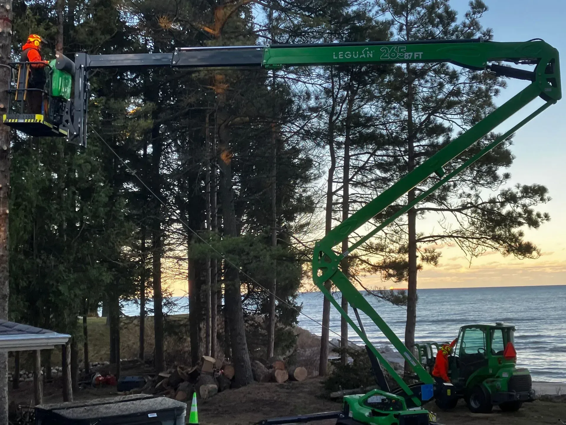 Green boom lift with two workers trimming tall trees near a lakeshore at sunset