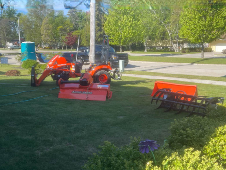 Orange lawn tractor with mower attachment on a grassy yard near a street, trees and houses in the background
