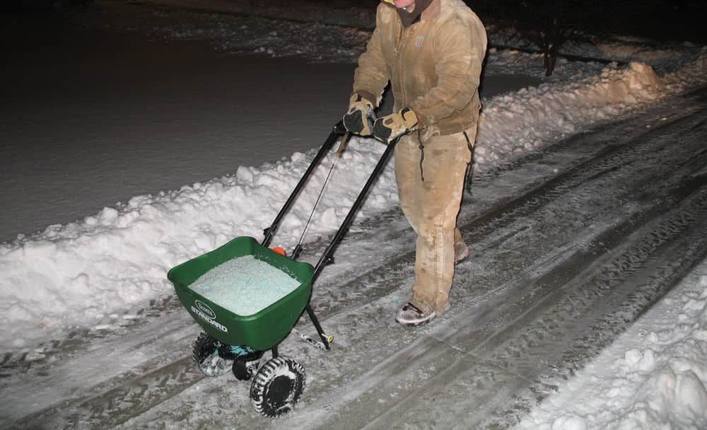 Person pushing a green spreader over a snowy driveway at night