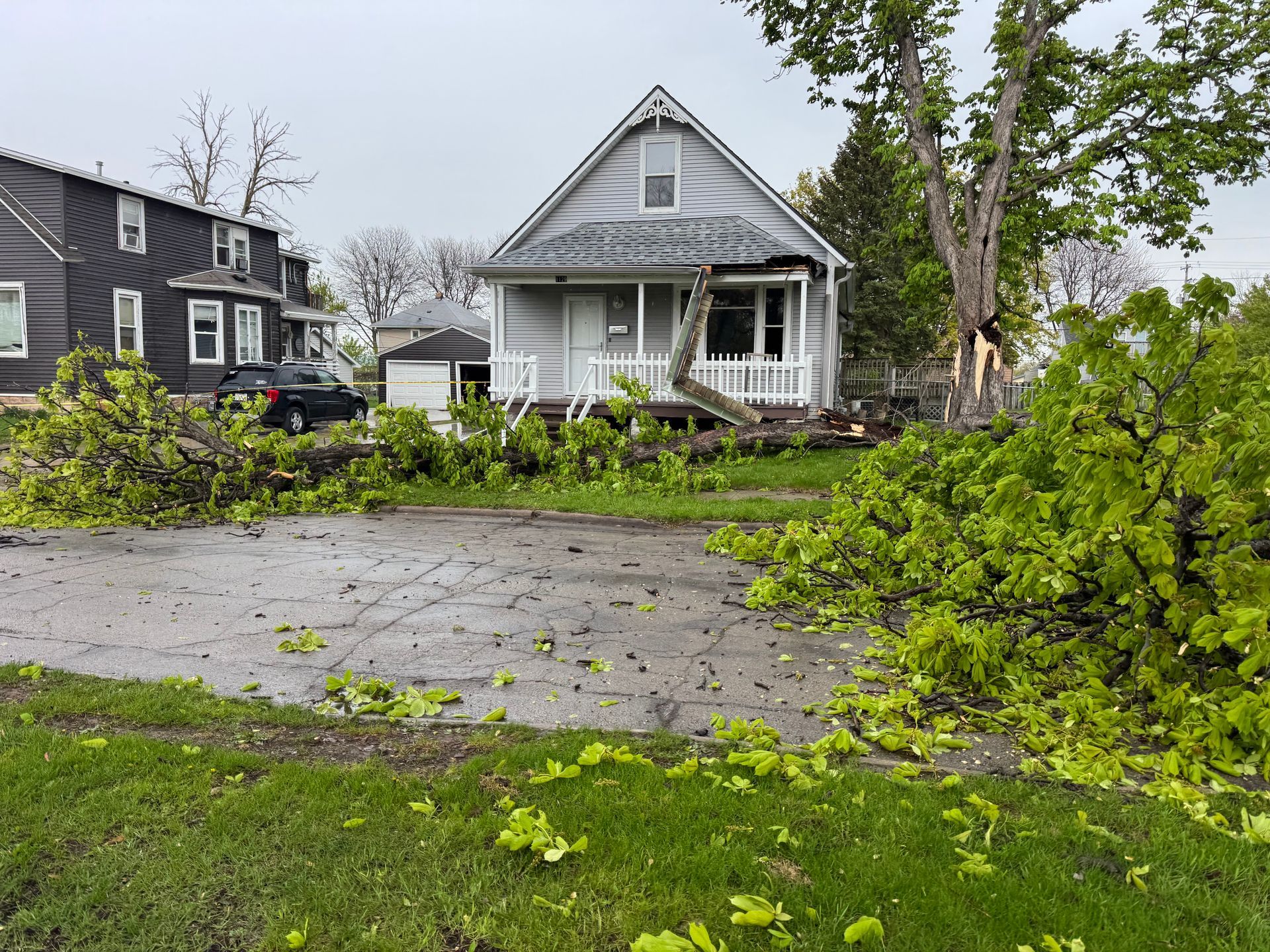 House and street blocked by storm debris, with fallen tree branches across the road and lawn.