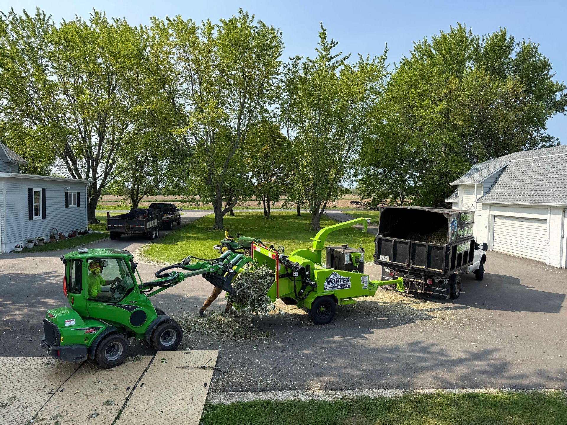 Green landscaping machines and a dump trailer parked in a driveway beside a garage and trees