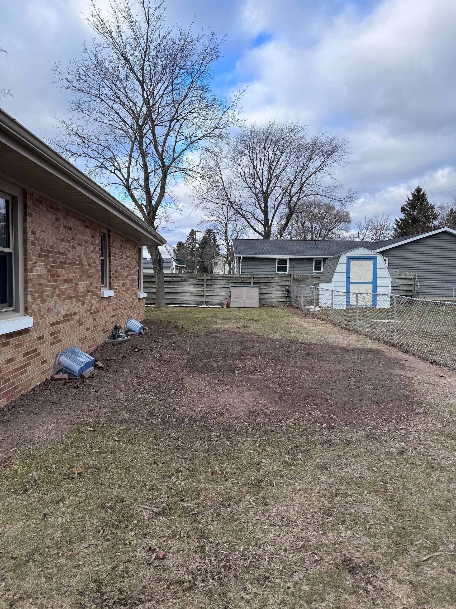 Backyard beside a brick house with bare trees, a brown lawn, fence, and a small white shed.