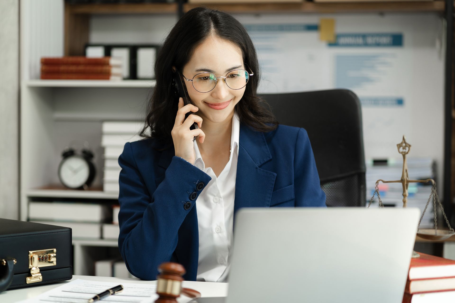 A woman is sitting at a desk talking on a cell phone in front of a laptop computer.