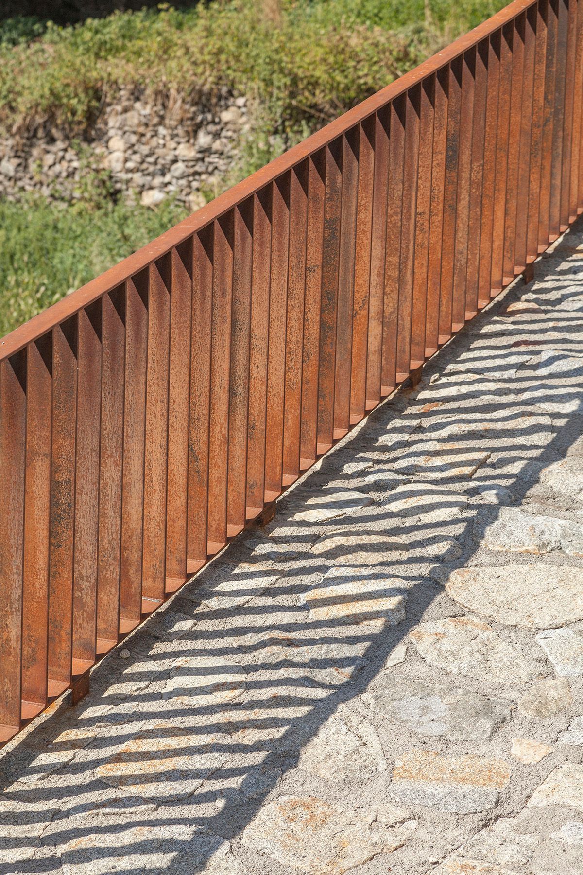 Una valla de metal oxidado proyecta una sombra sobre una pasarela de piedra. Emfasi Estudi