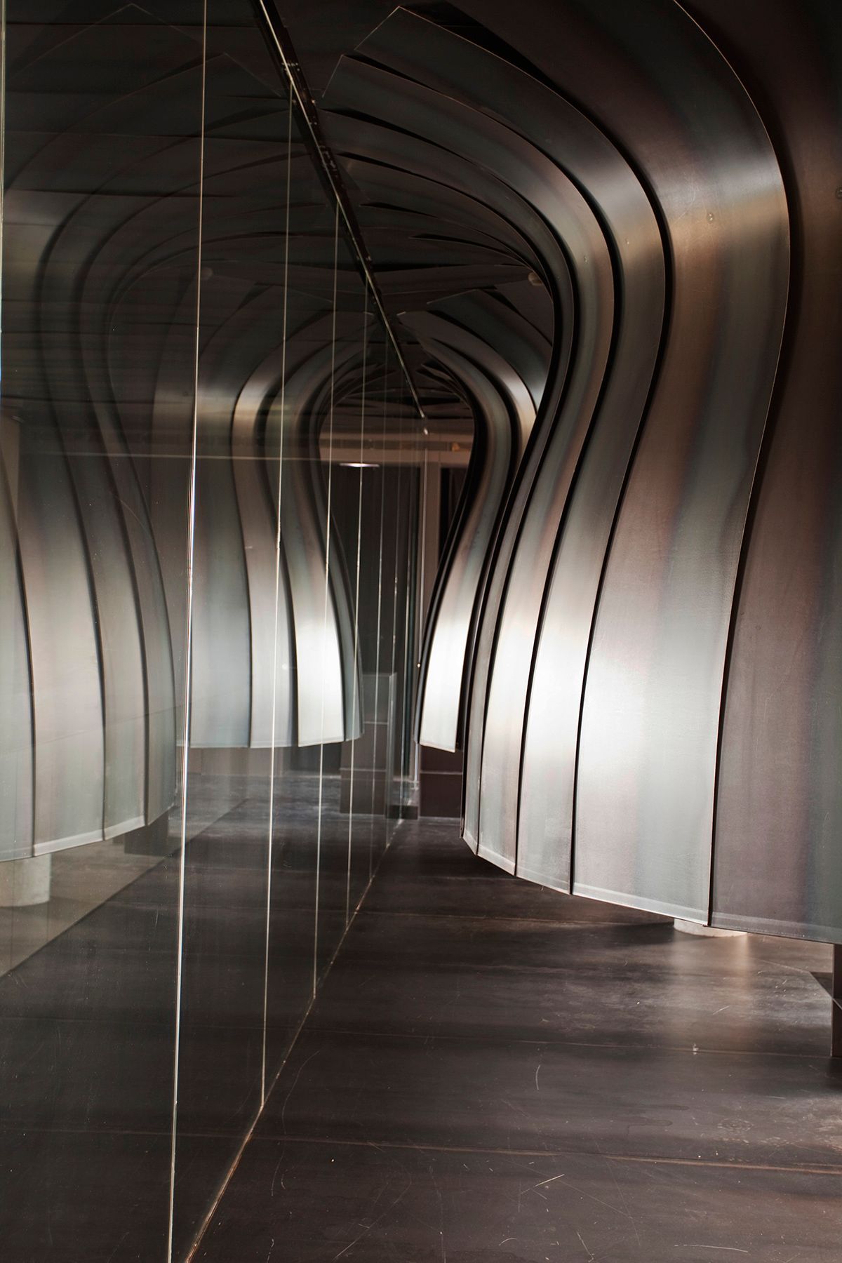 Fotografia Emfasi Estudi Andorra. Un pasillo largo con una pared metálica curva y suelo de madera. Arquitectura de vanguarda.