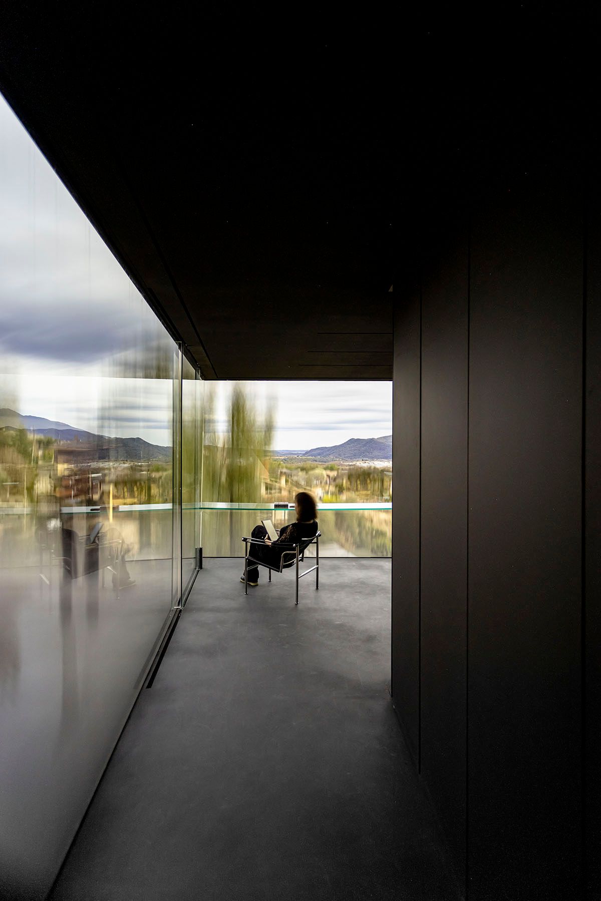 Fotografia Emfasi Estudi Andorra. Una persona está sentada en una silla en un balcón con vista a un lago. 