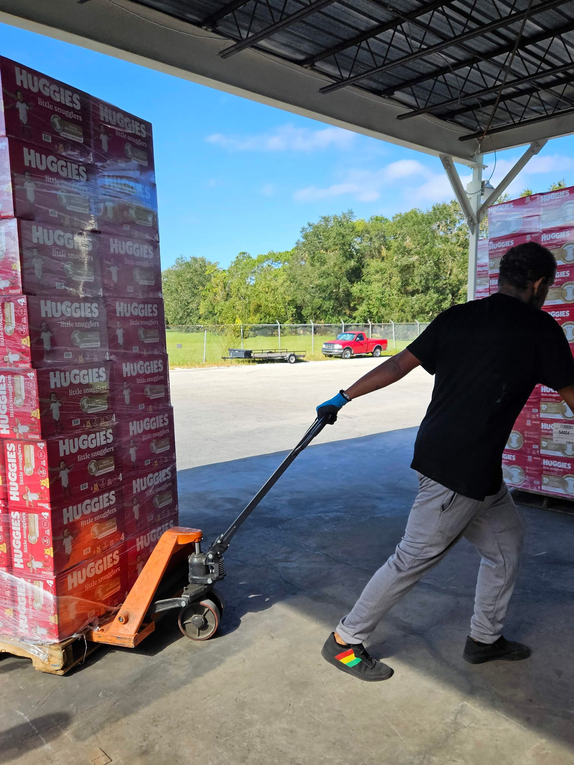 Person using a pallet jack to move stacked boxes under a covered loading area.