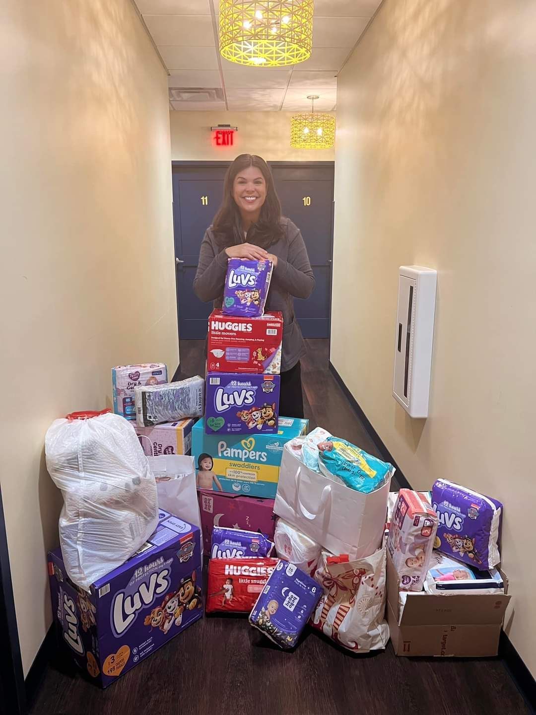 Person smiling in a hallway, surrounded by stacked boxes, bags, and packages of donated supplies