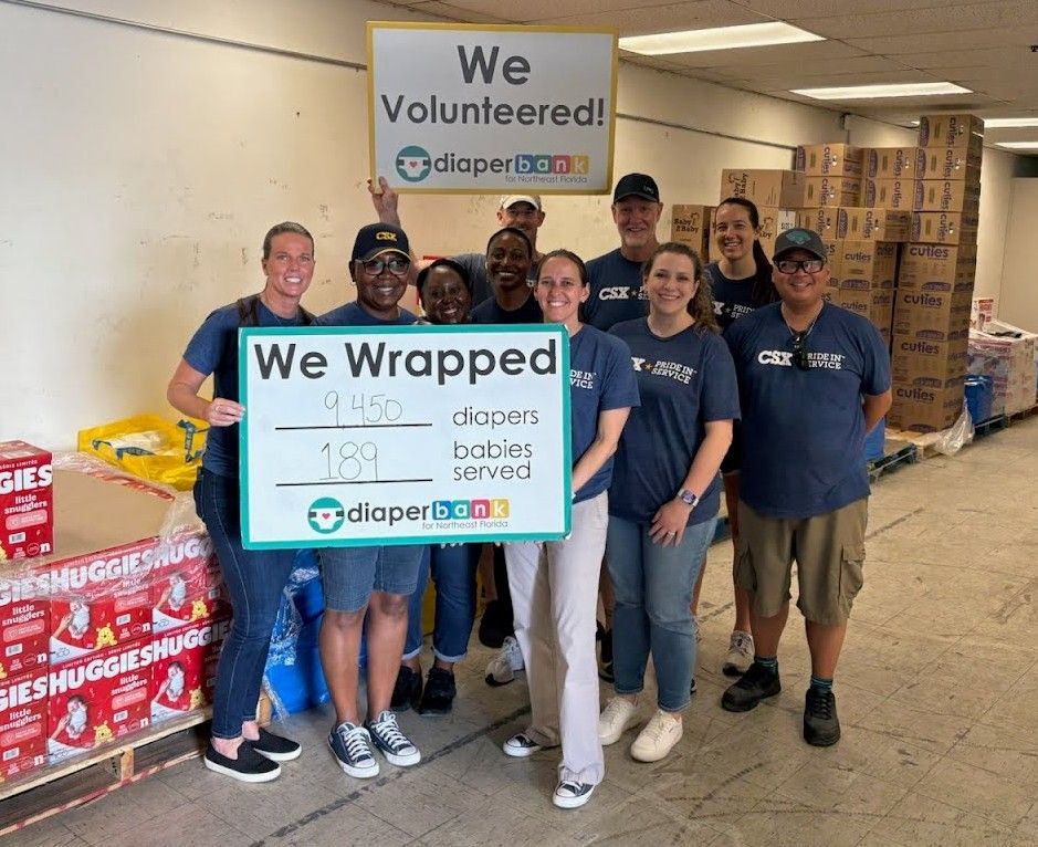 Volunteers holding “We Volunteered” and “We Wrapped” signs beside wrapped donations in a store.