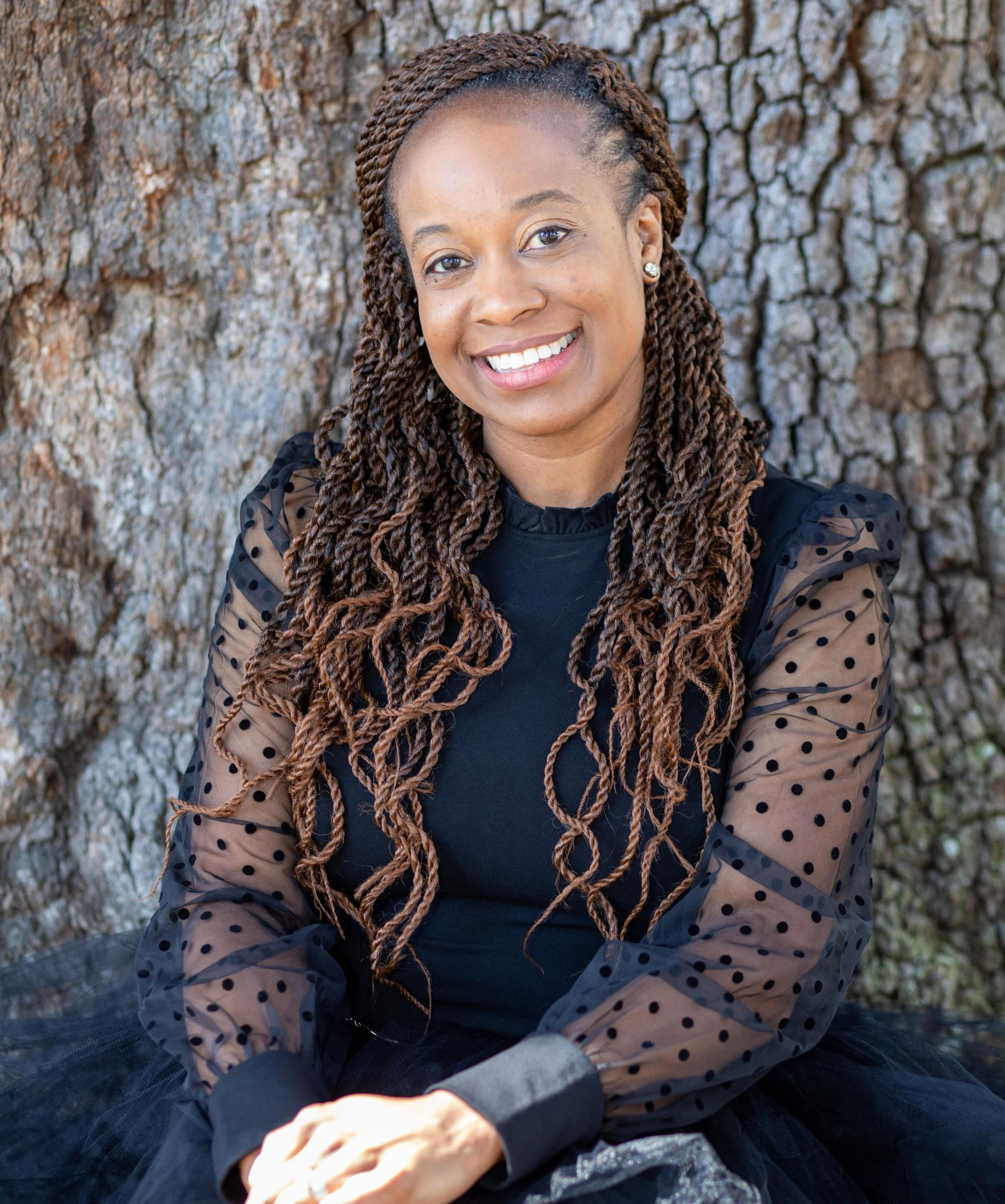 Smiling person in a black top sitting against a tree trunk outdoors