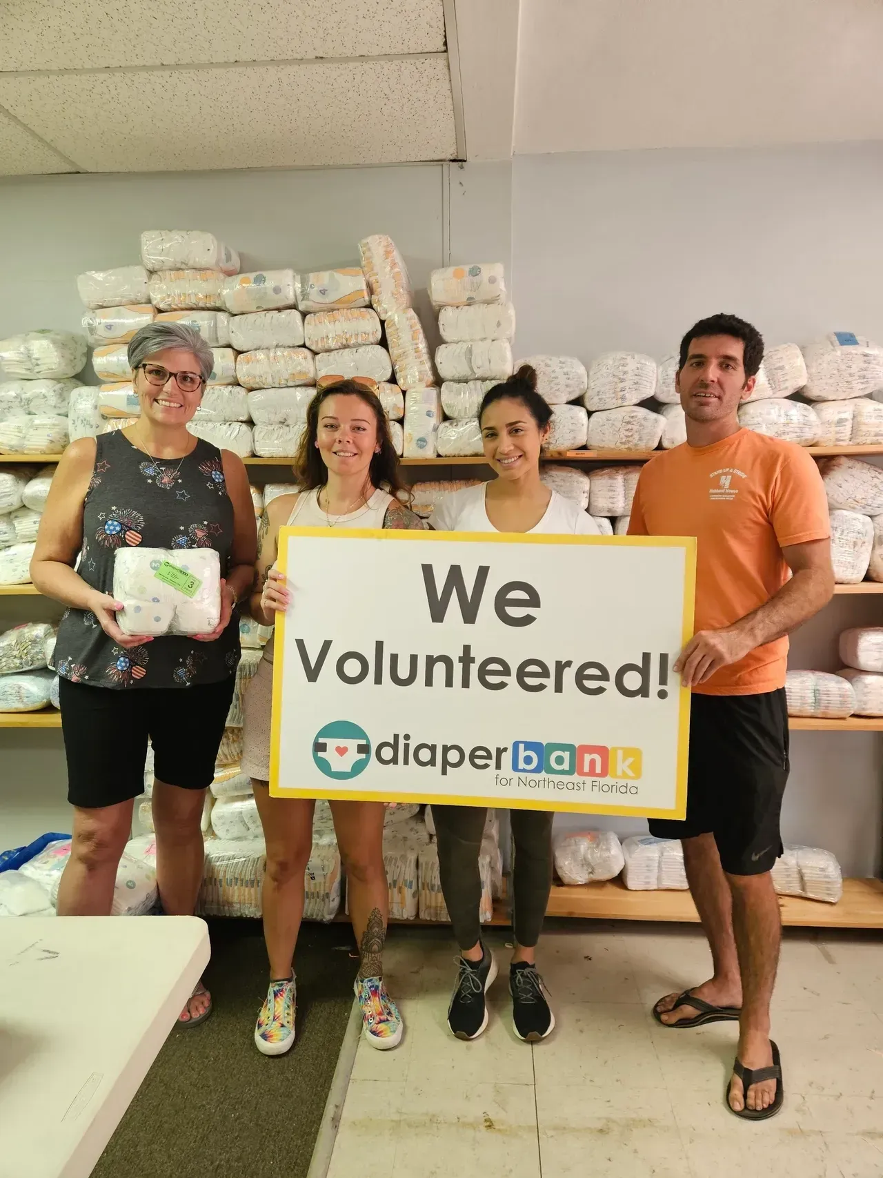 Four volunteers stand indoors holding a “We Volunteered!” banner in front of stacked supplies.