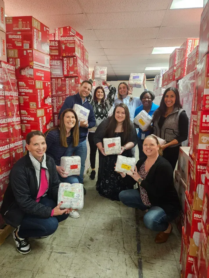 Group of volunteers in a warehouse aisle holding donated packages surrounded by stacked red boxes