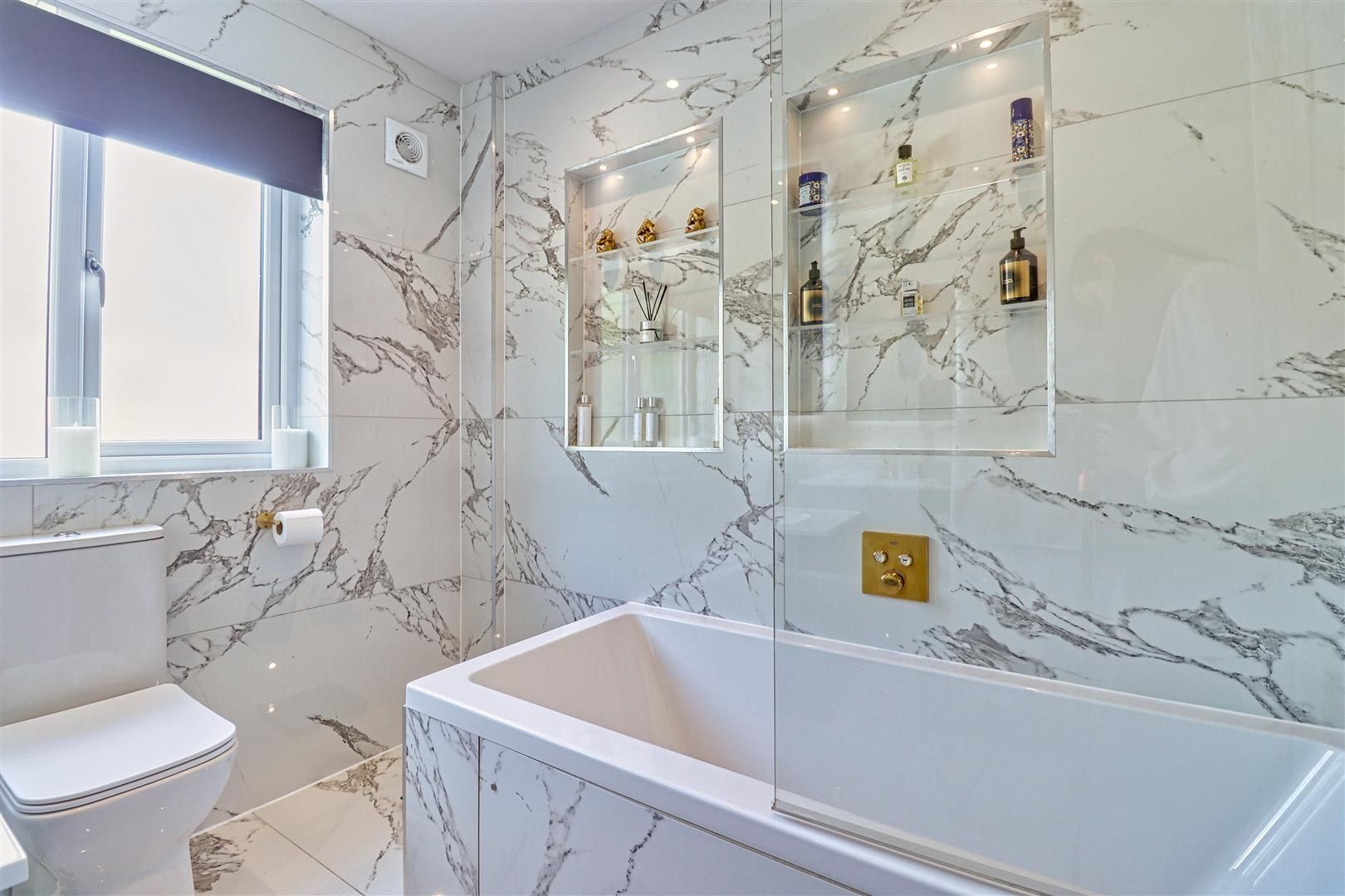 Marble-tiled bathroom with bathtub, toilet, recessed shelves with toiletries, and a window with a blind.