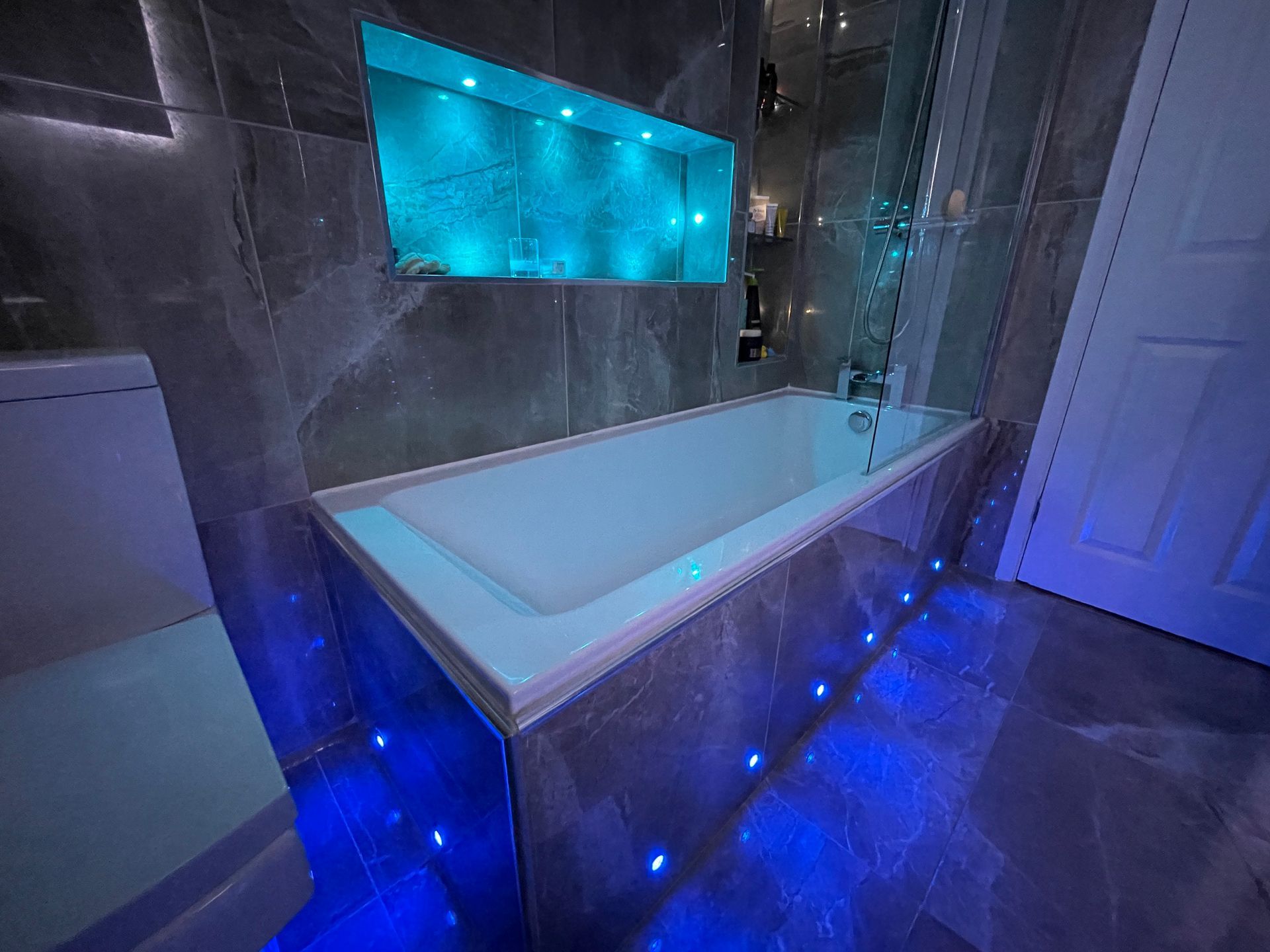 Bathroom with blue LED lights around the bathtub and in a recessed shelf, gray marble tile, and a white toilet.