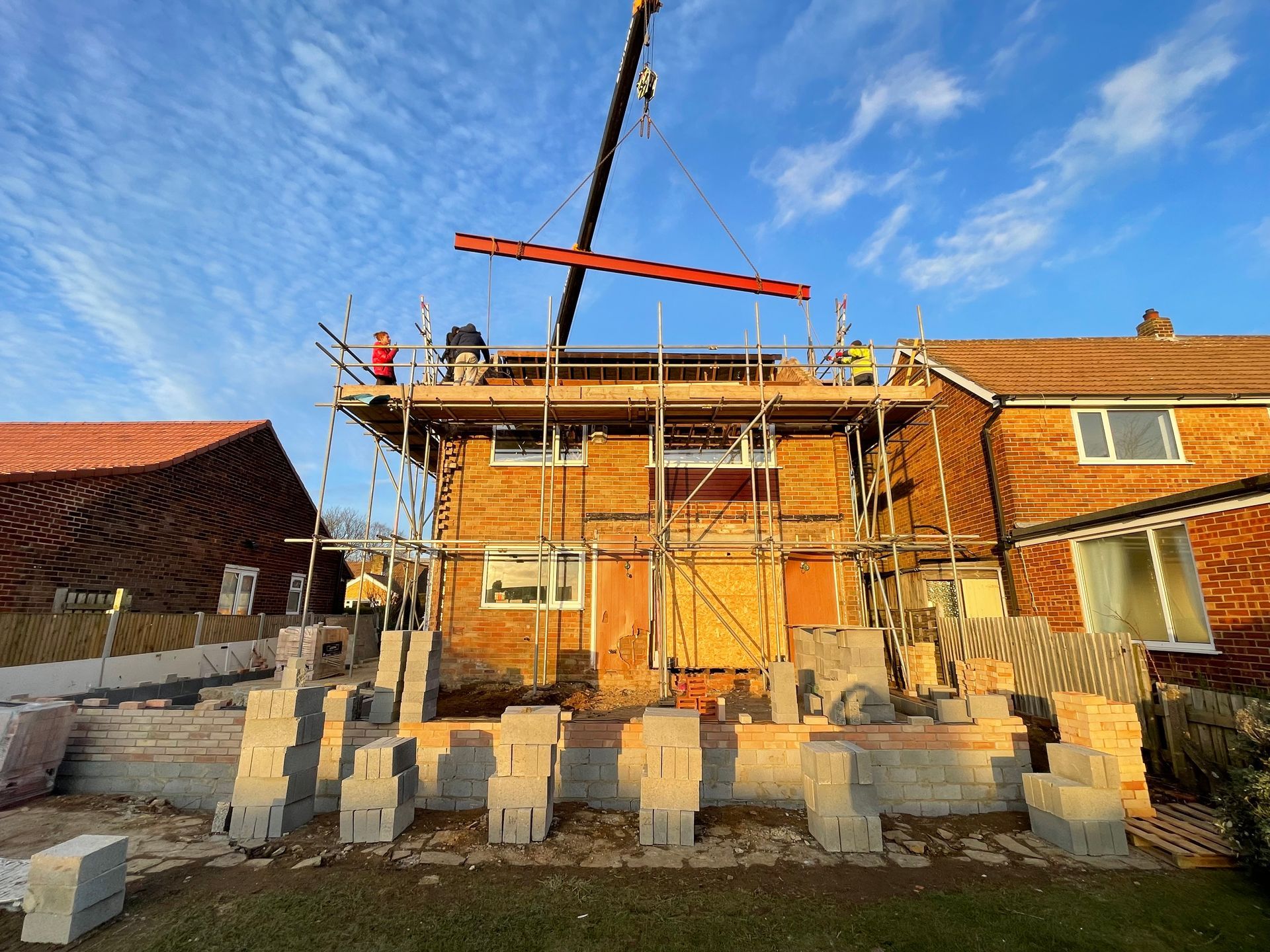 Construction of a brick house with scaffolding and a crane lifting a beam; sunny day.