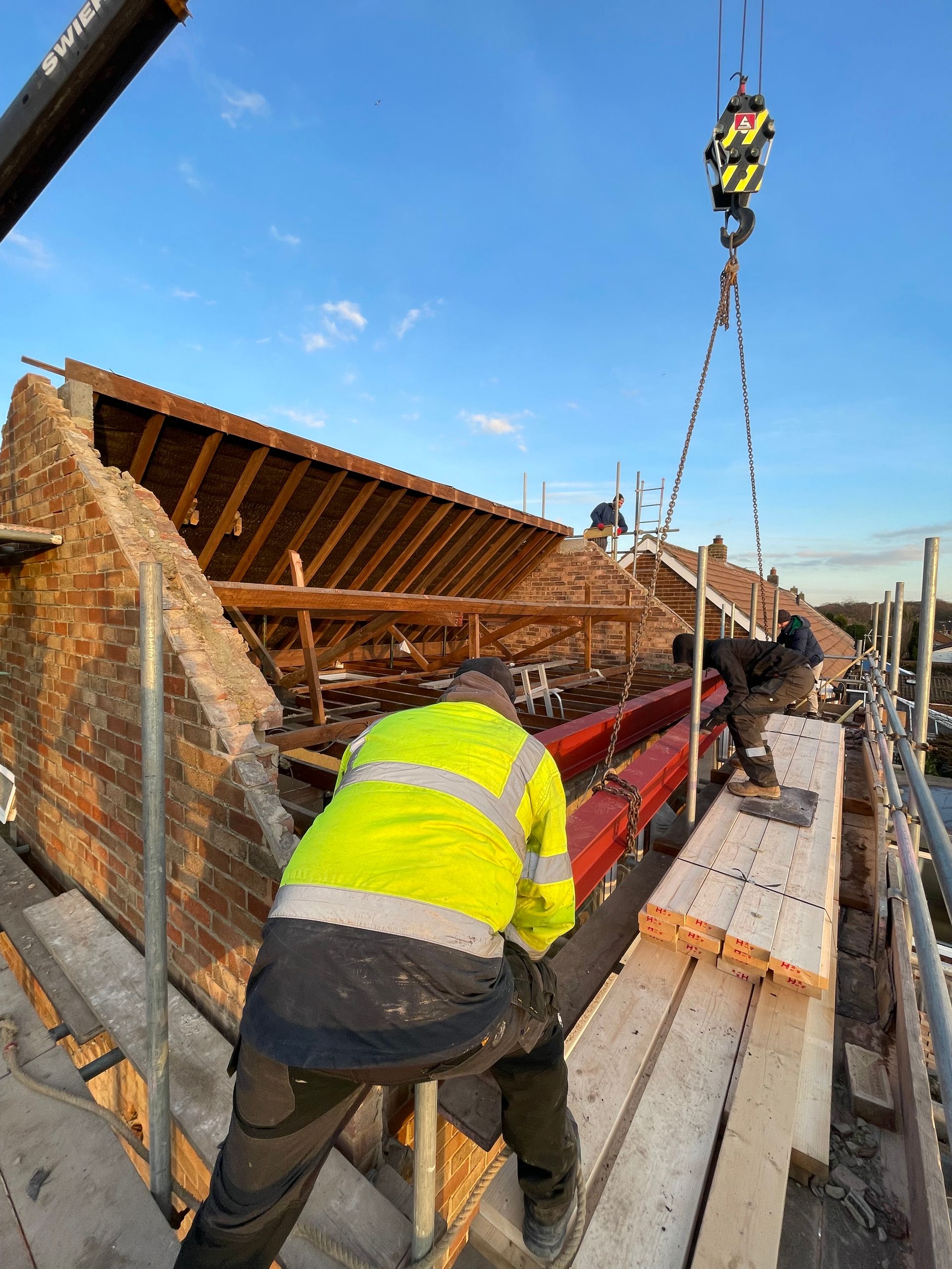 Construction workers on a roof using a crane to lift a red beam. Brick building with exposed rafters and scaffolding.